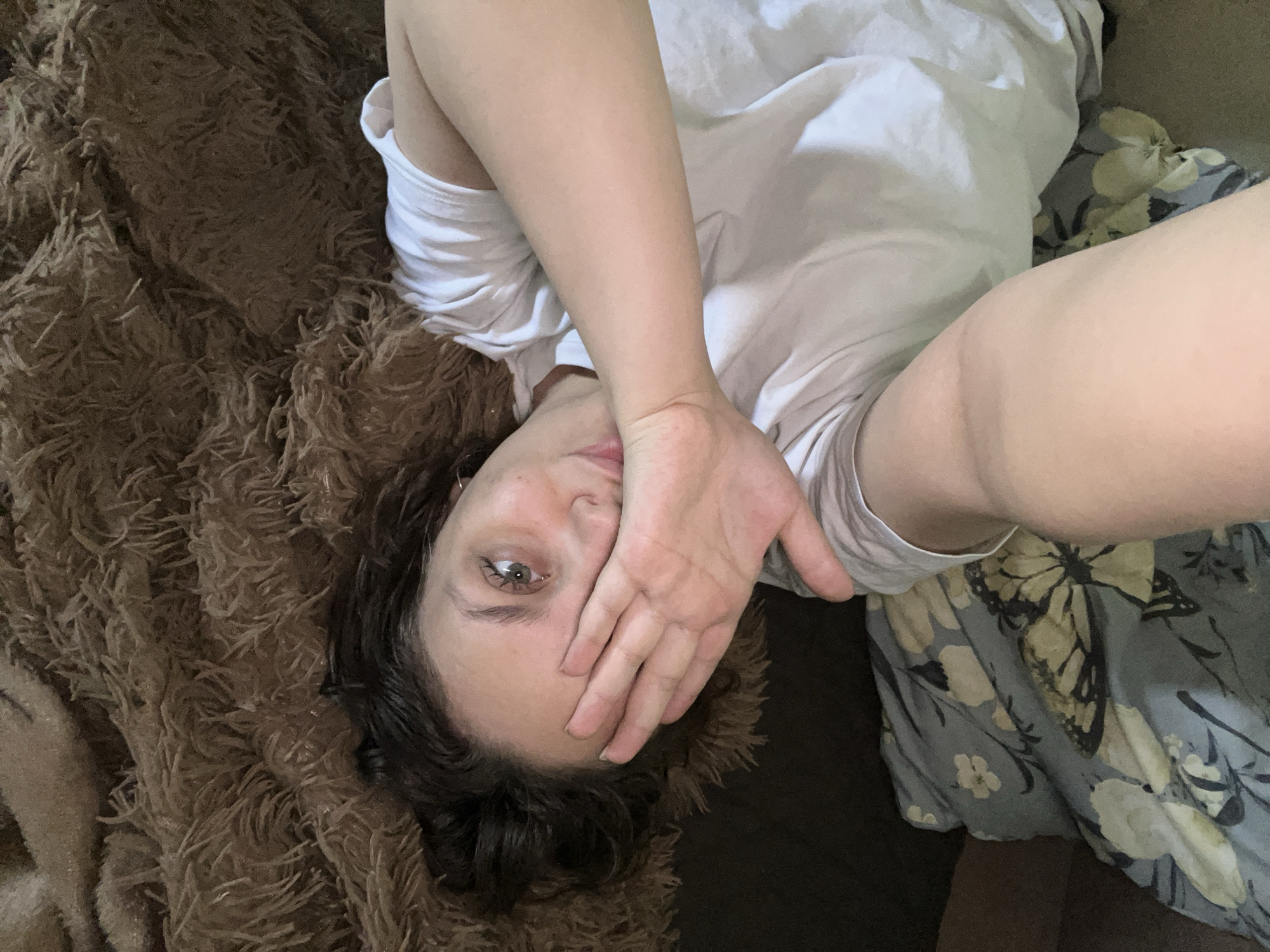A person is depicted in a casual, overhead shot, lying down indoors, likely on a bed or sofa. The individual, who appears to be young, has dark, wavy hair and fair skin. Their left hand covers the lower half of their face, from just below the nose, while their forehead and one distinct green eye are visible, gazing towards the camera. They are wearing a simple white short-sleeved t-shirt.
On the left side of the frame, there is a substantial amount of brown, shaggy or fluffy textured material, resembling a soft blanket or rug. On the right side, a contrasting fabric, possibly a pillowcase or another blanket, is visible. This fabric is blue-grey with a pattern of light-colored flowers and a prominent black and white butterfly, reminiscent of a monarch.
The person appears to be in a state of repose, possibly having just woken up or relaxing. The pose suggests a self-portrait or selfie, given the direct gaze into the lens and the partial facial obstruction often used in casual photography. The emotions conveyed are subtle, with the visible eye appearing calm or slightly contemplative. There are no specific external cues to indicate the time of day or weather. No visible text is present in the image. The setting is generic and does not offer specific location identifiers beyond being an indoor space.