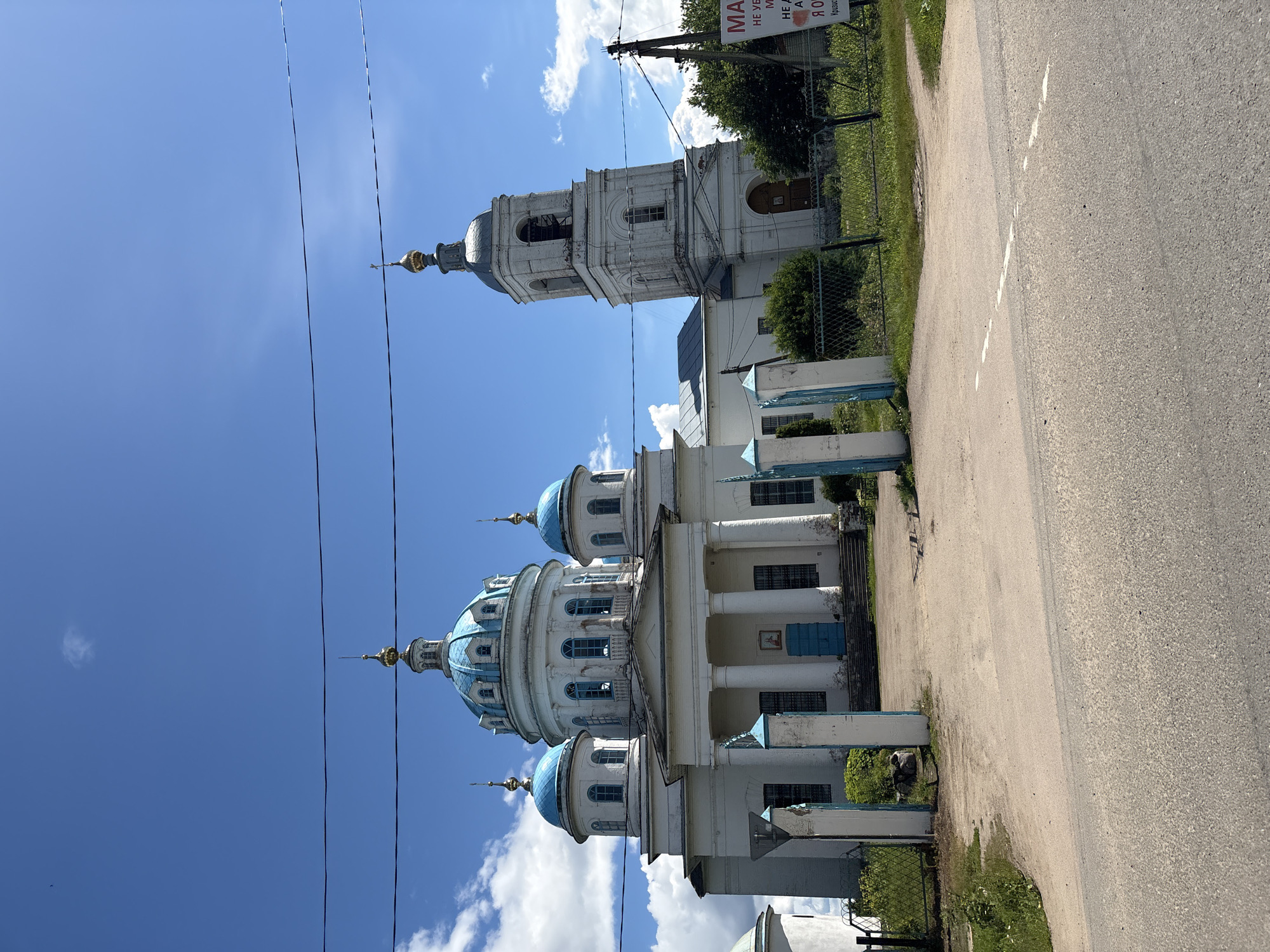 The image shows the Church of the Resurrection of Christ in Novoye, Russia. The church is a white building with a prominent blue-domed central structure and several smaller blue domes. To the right of the main building is a white bell tower with a grey dome. The church is surrounded by a fence and greenery. In the foreground, there is a paved road leading to an entrance gate with blue-topped pillars. The sky is blue with scattered white clouds. A sign is visible on the right side of the image with Russian text. No people are present in the image.