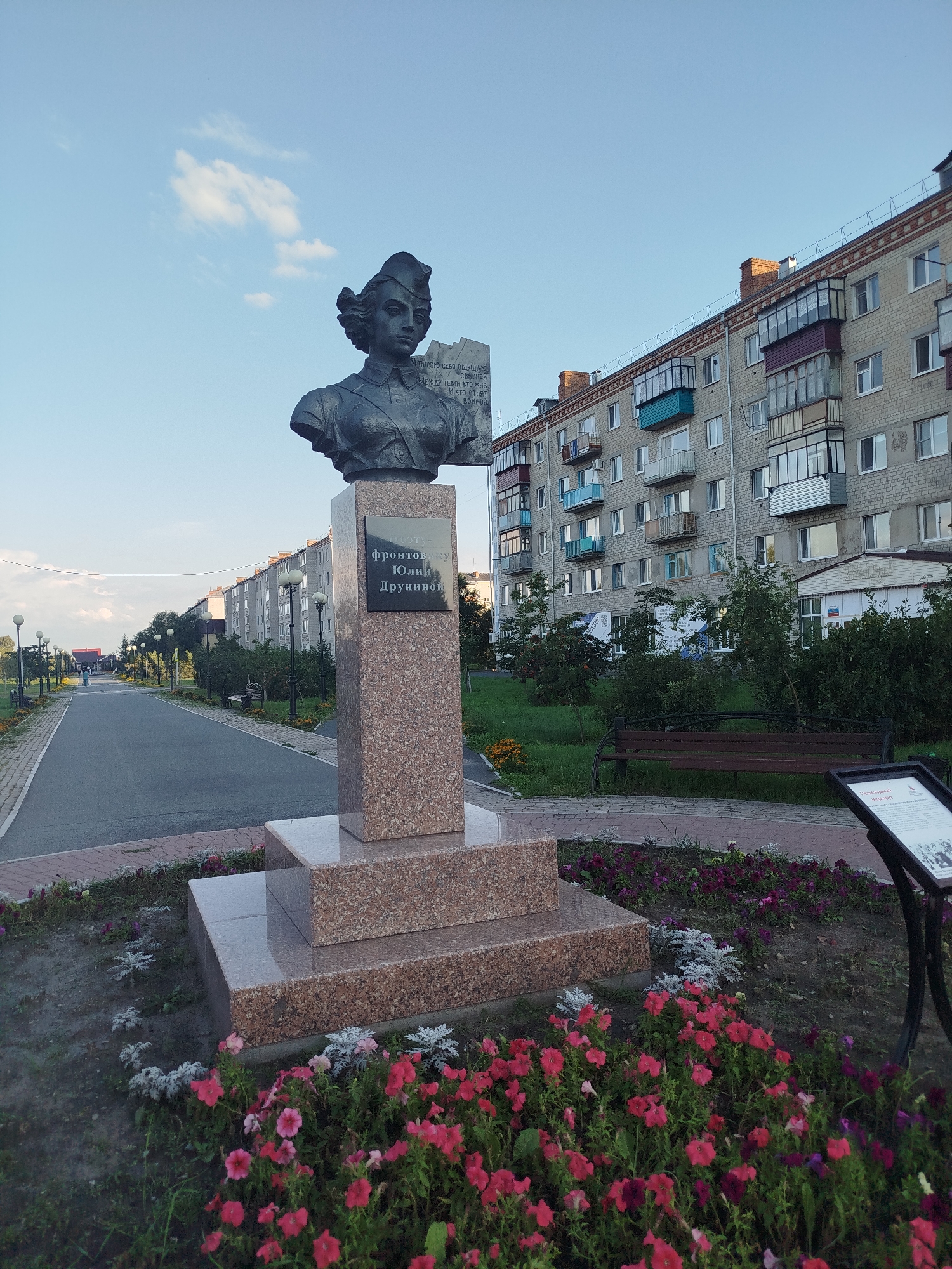 This is a full-frame, eye-level, outdoor photograph taken in the city of Yalutorovsk, Russia. The image features a bronze bust of a woman in a military cap and uniform, standing on a multi-tiered granite pedestal. The bust is positioned on the right side of the frame. A plaque on the pedestal is inscribed with Russian text. In the background, a long, multi-story apartment building stretches across the frame, with visible balconies and windows. Adjacent to the building is a park area with benches, trees, and flowerbeds. A paved walkway leads away from the foreground into the distance, lined with lampposts and trees. The sky is clear with a few scattered clouds. The foreground is dominated by a vibrant flowerbed filled with pink and purple flowers.