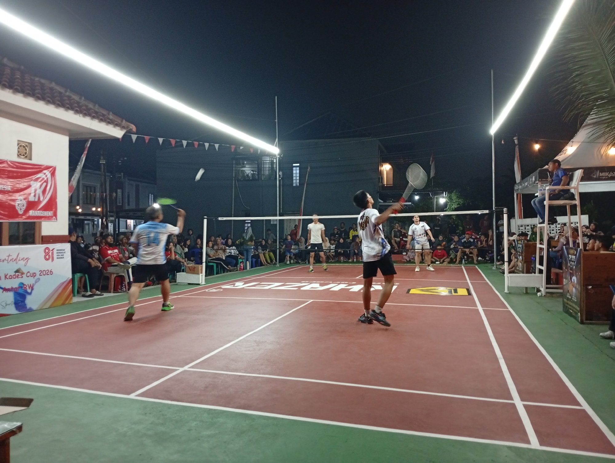 The image depicts an outdoor badminton match at night on a red court with green borders, illuminated by bright overhead lights. Spectators are seated on benches and chairs along the side of the court. Several players are visible on the court, actively engaged in the game, with one player in the foreground mid-swing. Banners and flags are displayed, including one that reads "Kades Cup 2026." The background shows buildings and trees, suggesting an urban or semi-urban setting in Caringin, Indonesia. The overall atmosphere appears to be that of a local sporting event.