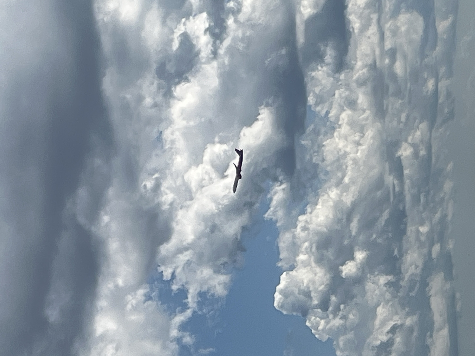 An airplane is visible in the sky, flying through a cloudy sky with patches of blue. The airplane is silhouetted against the clouds and is angled slightly upwards, suggesting it is ascending or cruising. The clouds are varied, with some dark and heavy-looking, and others light and fluffy. The overall impression is of a dynamic and somewhat overcast sky. There is no identifiable location in Varna, Bulgaria.