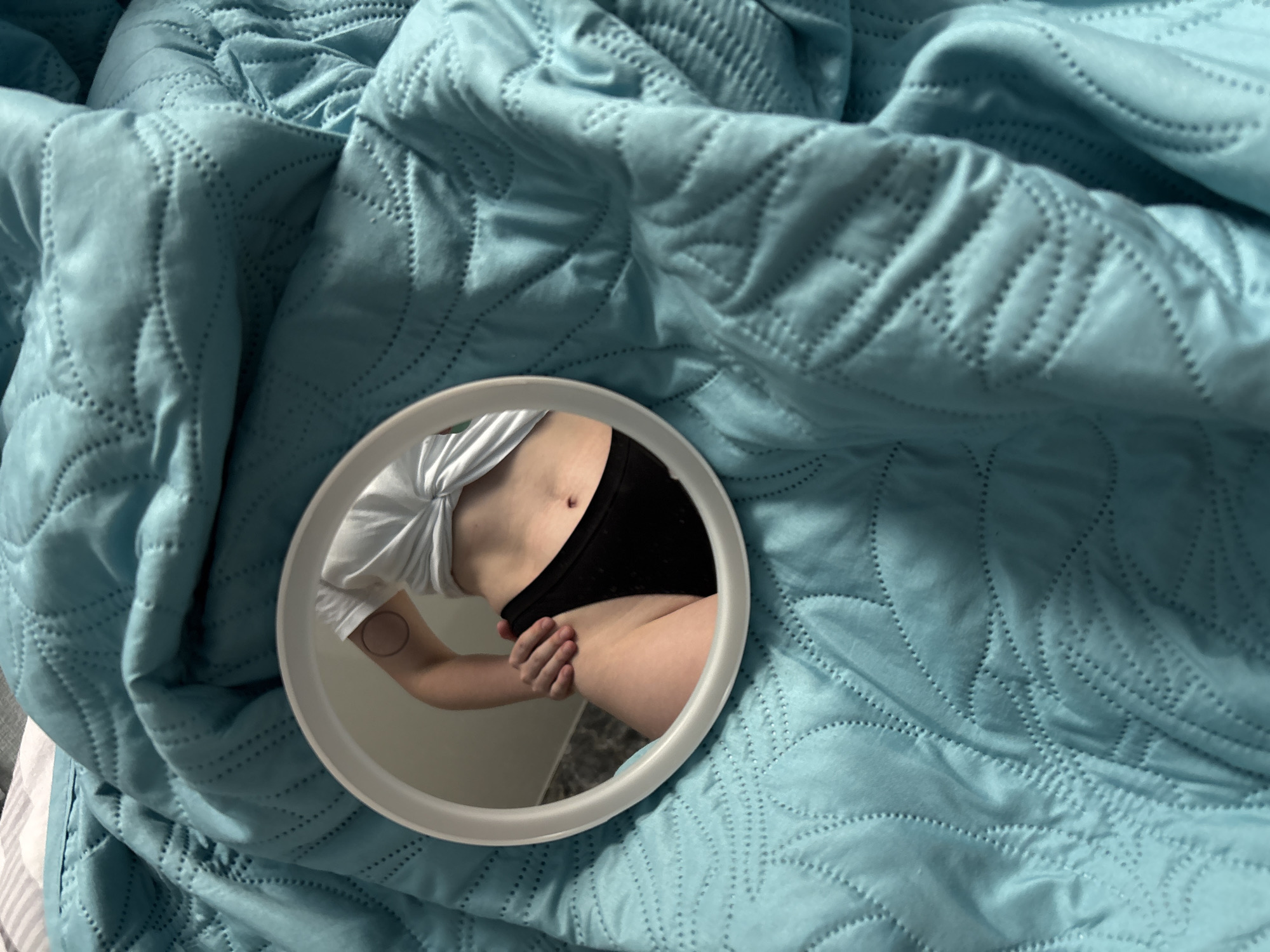 A round mirror reflects a person's torso and arm, resting on a quilted, teal blanket. The person is wearing a white, knotted t-shirt and black underwear. A small, circular tattoo is visible on their upper arm. The blanket has a diamond-shaped pattern created by stitching. The lighting suggests it is daytime. The reflection shows the person's midriff, and they are holding their hip with their left hand. The background in the reflection appears to be a wall. The scene is likely indoors, possibly a bedroom. There is no visible text in the image.