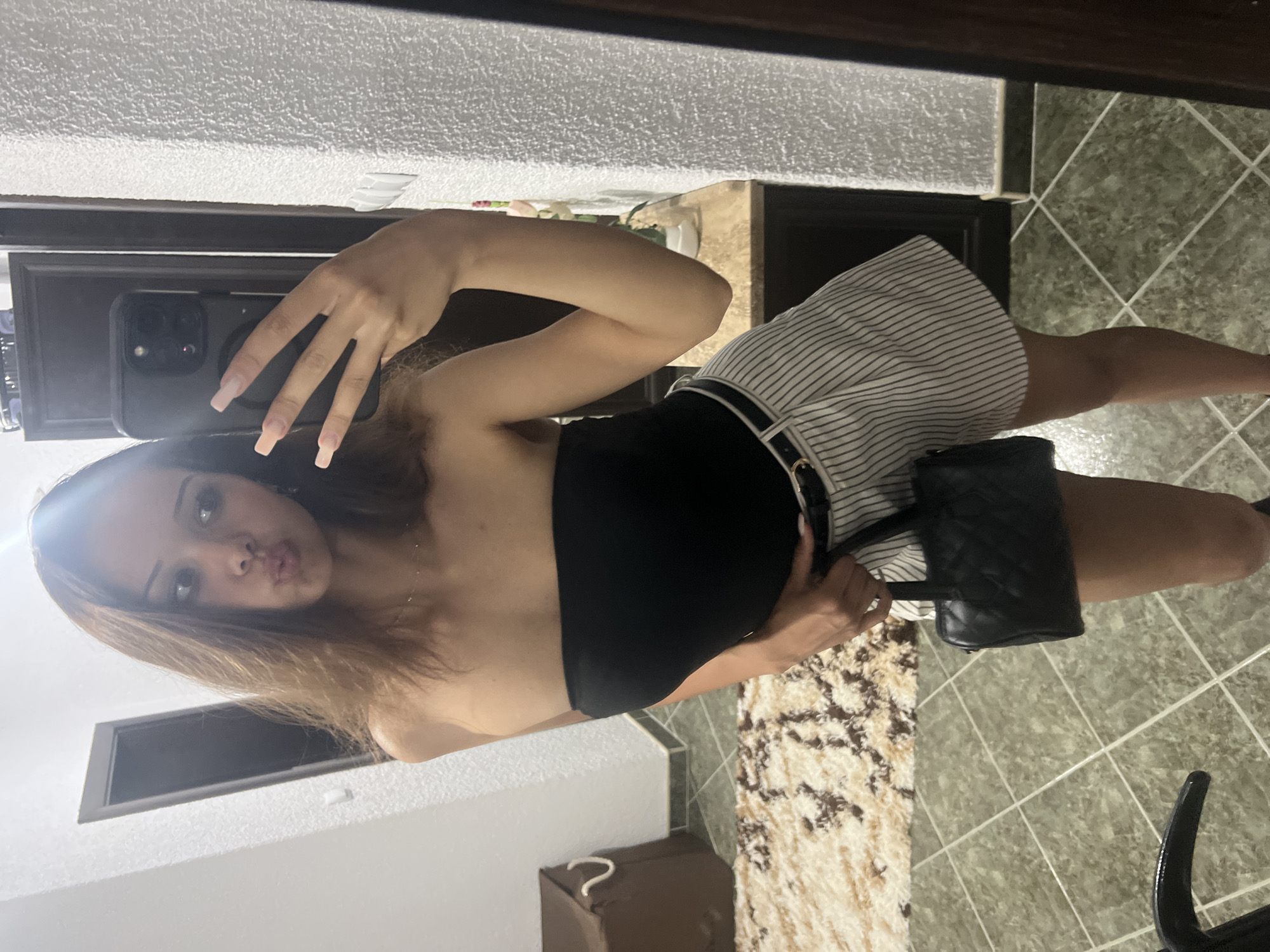 A young woman is taking a selfie in a room. She is wearing a black strapless top and white and black striped shorts. She is holding a black quilted handbag. The room has tiled floors and a textured wall. There is a rug on the floor and a laundry basket in the background. The woman is leaning against a wall and a door is visible behind her. The lighting is dim.