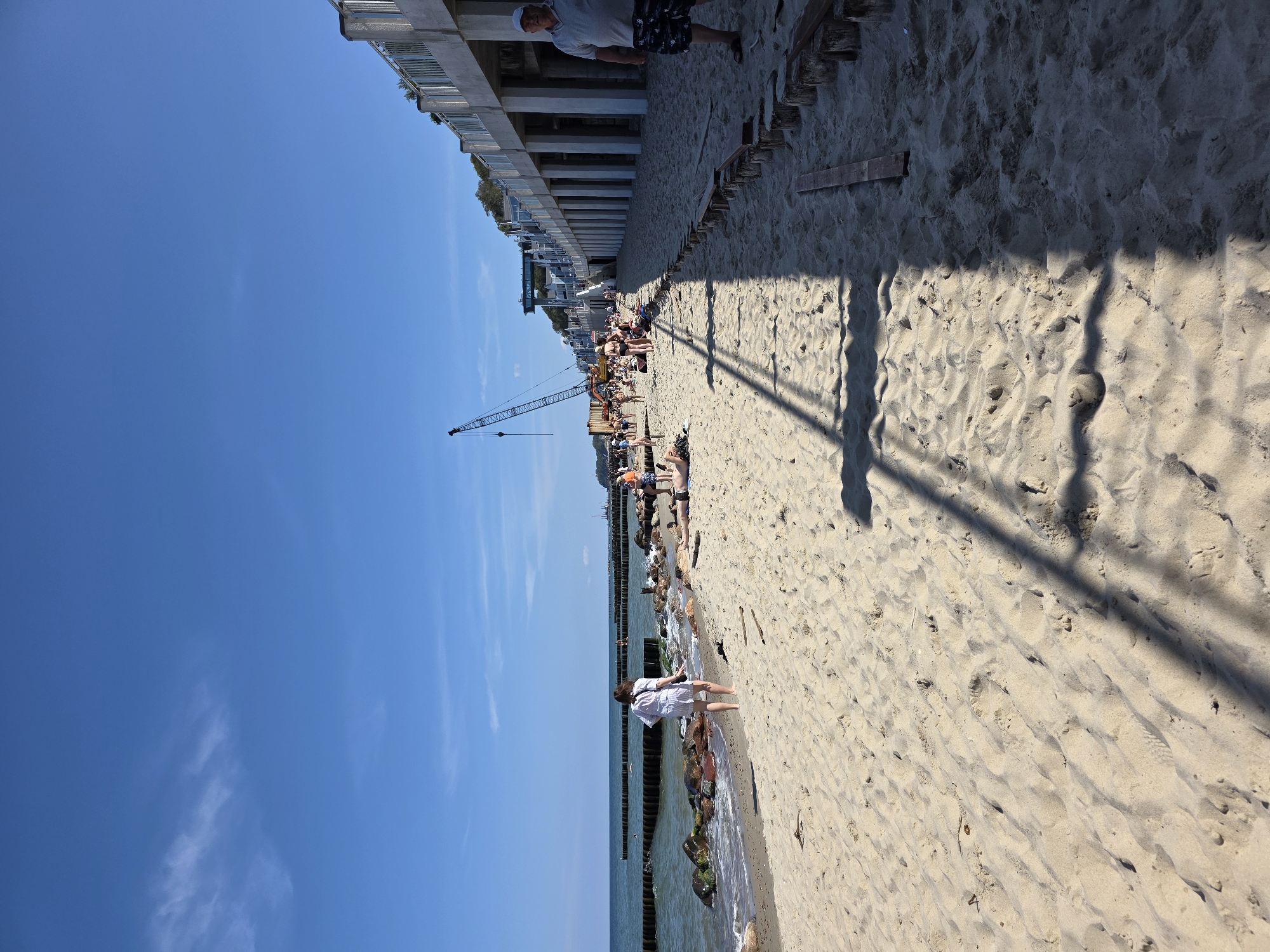 A sunny day at Svetlogorsk beach in Russia, with clear blue skies and a few wispy clouds. The sandy shore is populated by beachgoers enjoying the weather. In the background, a large crane is visible, suggesting ongoing construction or maintenance work on the coastal infrastructure. A raised walkway or promenade runs along the edge of the beach, supported by pillars, with people strolling along it. The sea stretches out to the horizon, with wooden breakwaters dotting the water.