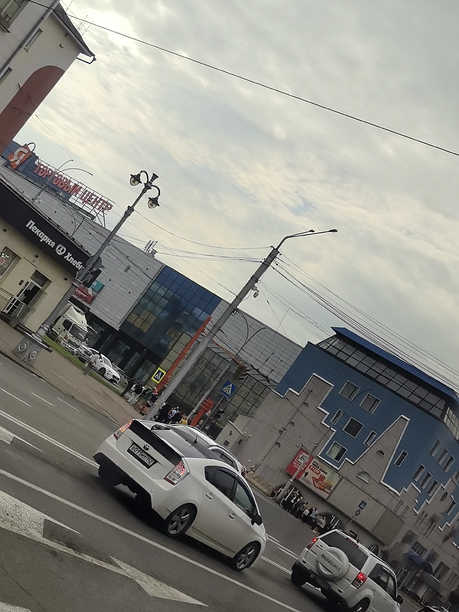 The image depicts a street scene in an urban environment, likely in Russia, given the Cyrillic text visible on signage. The viewpoint is from a vehicle traveling on a road. The sky is overcast with a moderate amount of clouds.
Prominently featured is a modern, multi-story building with a distinctive blue facade, featuring a unique, geometric architectural design with numerous windows. To the left of this building, another structure with a light-colored facade and a red arched entrance is visible. Signage on this building indicates "ТОРГОВЫЙ ЦЕНТР" (Shopping Center) and "Пекарня @ Хлеби" (Bakery @ Bread).
Several vehicles are present on the road and parked. Two white cars are the most visible: a Suzuki Vitara with a spare tire mounted on the rear, and a Toyota Prius with a visible license plate "B011MT142". A white van or small truck is parked near the bakery.
A group of pedestrians is gathered on the sidewalk in front of the blue building, some appearing to be waiting. Streetlights and traffic signals are also visible. The road has clear lane markings, including directional arrows. The overall impression is a busy commercial area on a cloudy day.