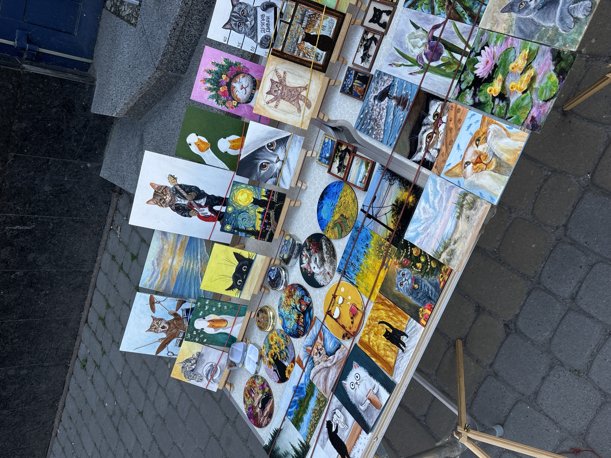 The image displays an outdoor art stall featuring a variety of paintings, primarily of cats in different styles and settings, along with some nature scenes. The stall is set up on a cobblestone pavement, with a dark, textured wall and a stone staircase visible in the background. The art pieces are small and displayed on miniature easels, secured with red elastic bands. Some of the paintings depict cats playing musical instruments, wearing hats, or interacting with natural elements like flowers and water. There are also circular artworks and small framed miniatures. The lighting suggests it is daytime, possibly late afternoon. The general setting appears to be a pedestrian area, potentially a market or a promenade in the city of Ternopil, Ukraine. No people are visible in the frame.