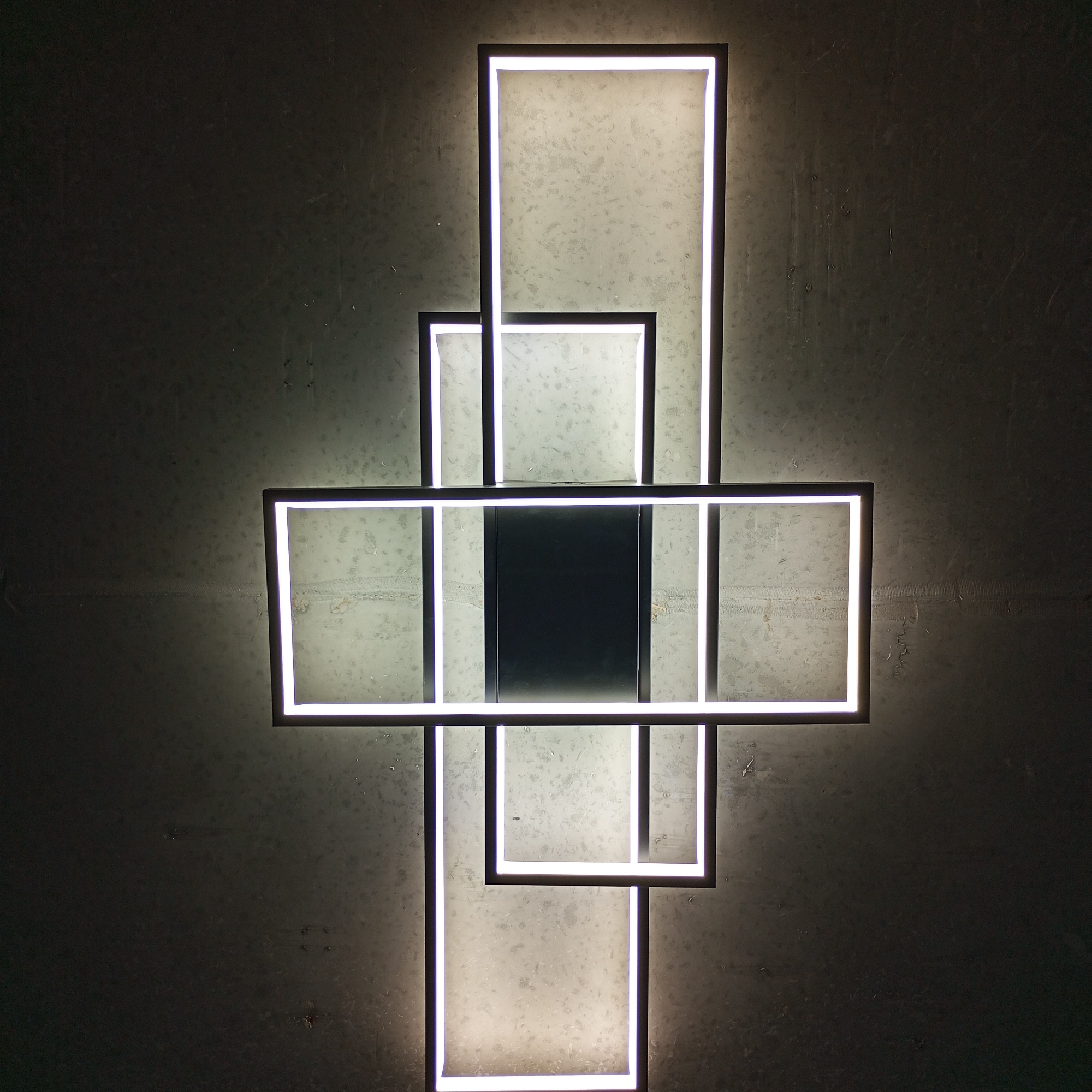 The image shows a modern, abstract wall-mounted light fixture. It consists of a series of interconnected rectangular frames of varying sizes. The frames are dark gray or black, with bright white light emanating from the inside edges, creating a linear illumination effect. The arrangement is vertically oriented and resembles a stylized cross or an elongated plus sign.
The fixture is mounted on a gray or concrete wall with a textured or mottled surface. The overall lighting is dim, focusing attention on the illuminated light fixture.
There is no evidence that this image is located in the city of Pervomaysky, Russia.