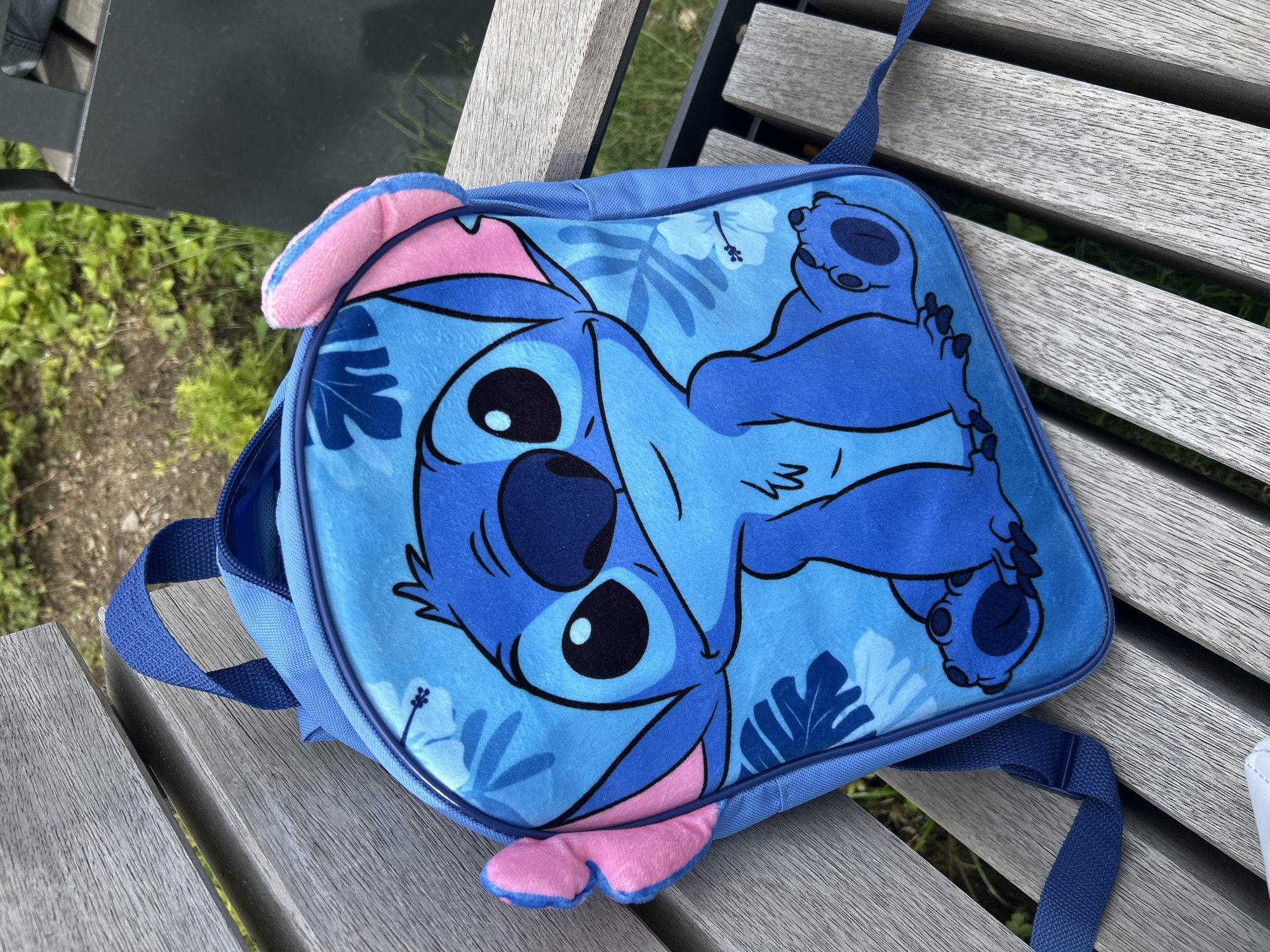 A blue children's backpack featuring the character Stitch from Disney's Lilo & Stitch is placed on a weathered wooden bench. The backpack has a plush texture and displays a large image of Stitch with his characteristic large eyes and ears. The character is depicted in a sitting pose with a tropical floral pattern in shades of blue and white in the background. The backpack has two adjustable blue straps. The bench is outdoors, with blurred green foliage and a dark object visible in the background, suggesting an outdoor setting such as a park or public seating area. The location is the city of Waldbröl, Germany.
