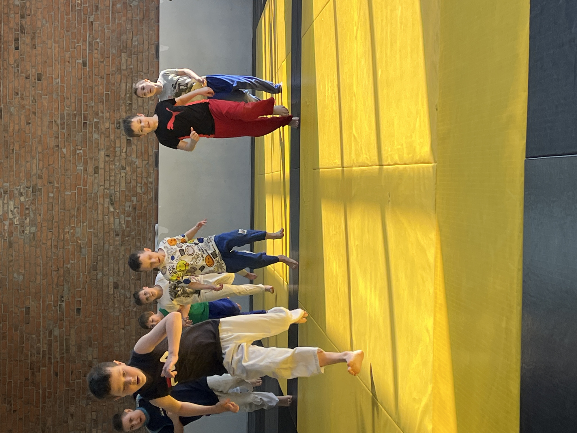 A group of seven children are standing on a yellow mat with black edges in what appears to be a room for martial arts or similar physical activity, evidenced by the mat. The children are of varying ages and are wearing casual clothing, including T-shirts and pants in various colors. The background consists of a brick wall on one side and a gray wall on the other. The lighting appears to be natural, with some shadows cast by the children. There are no indications that this image was taken in the city of Krasnoyarsk, Russia.