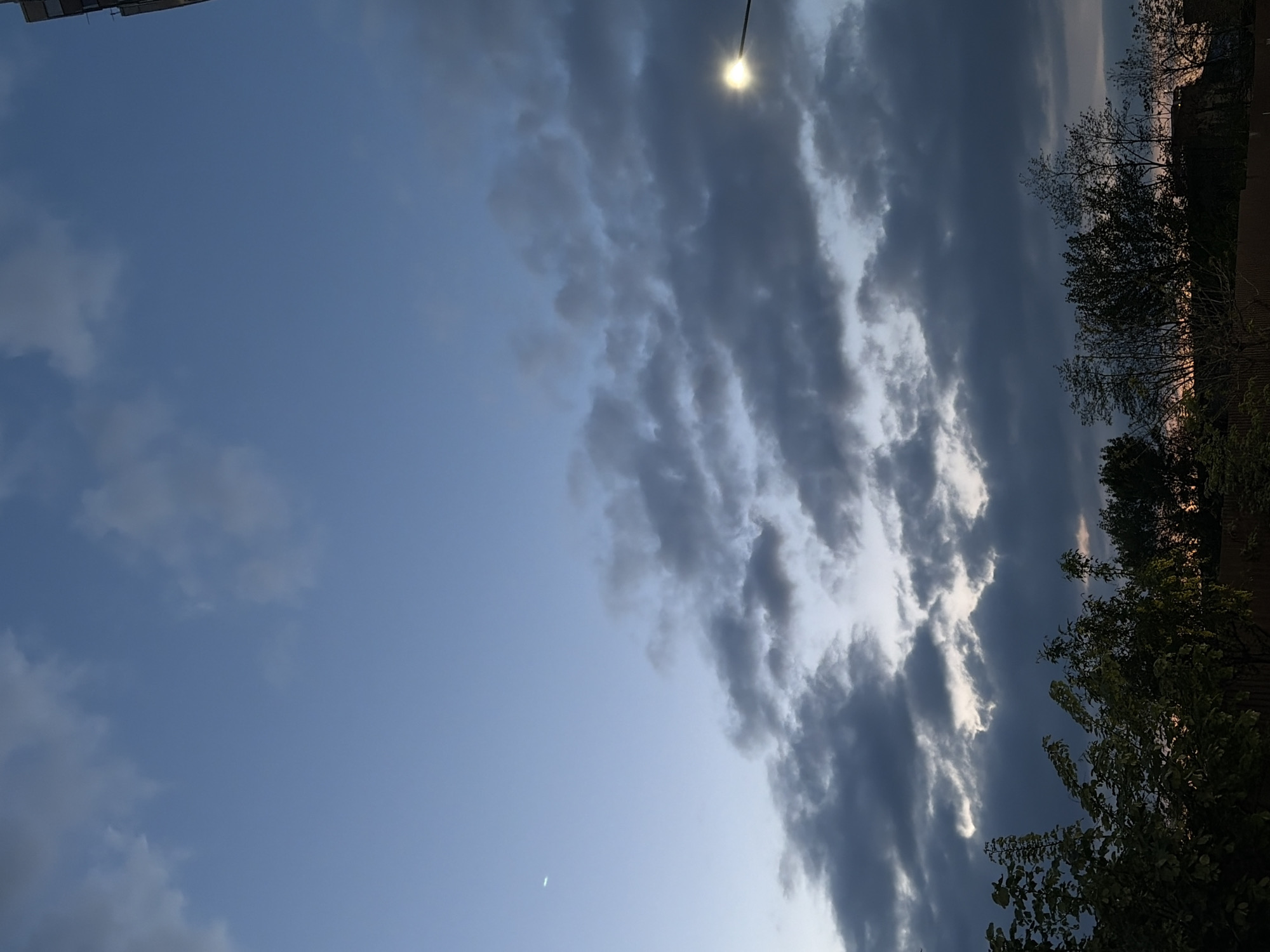 The image shows a sky scene, likely in the evening or at dusk, given the lighting. The sky is partially clear with blue hues on the left side and a significant band of dark, dense clouds stretching diagonally across the frame. A street light is visible near the top of the frame, emitting a bright glow, suggesting it is turned on. Along the right edge, trees are silhouetted against the sky. A thin crescent moon is visible. The scene appears to be from an urban or suburban location, and is in Krasnodar, Russia.
