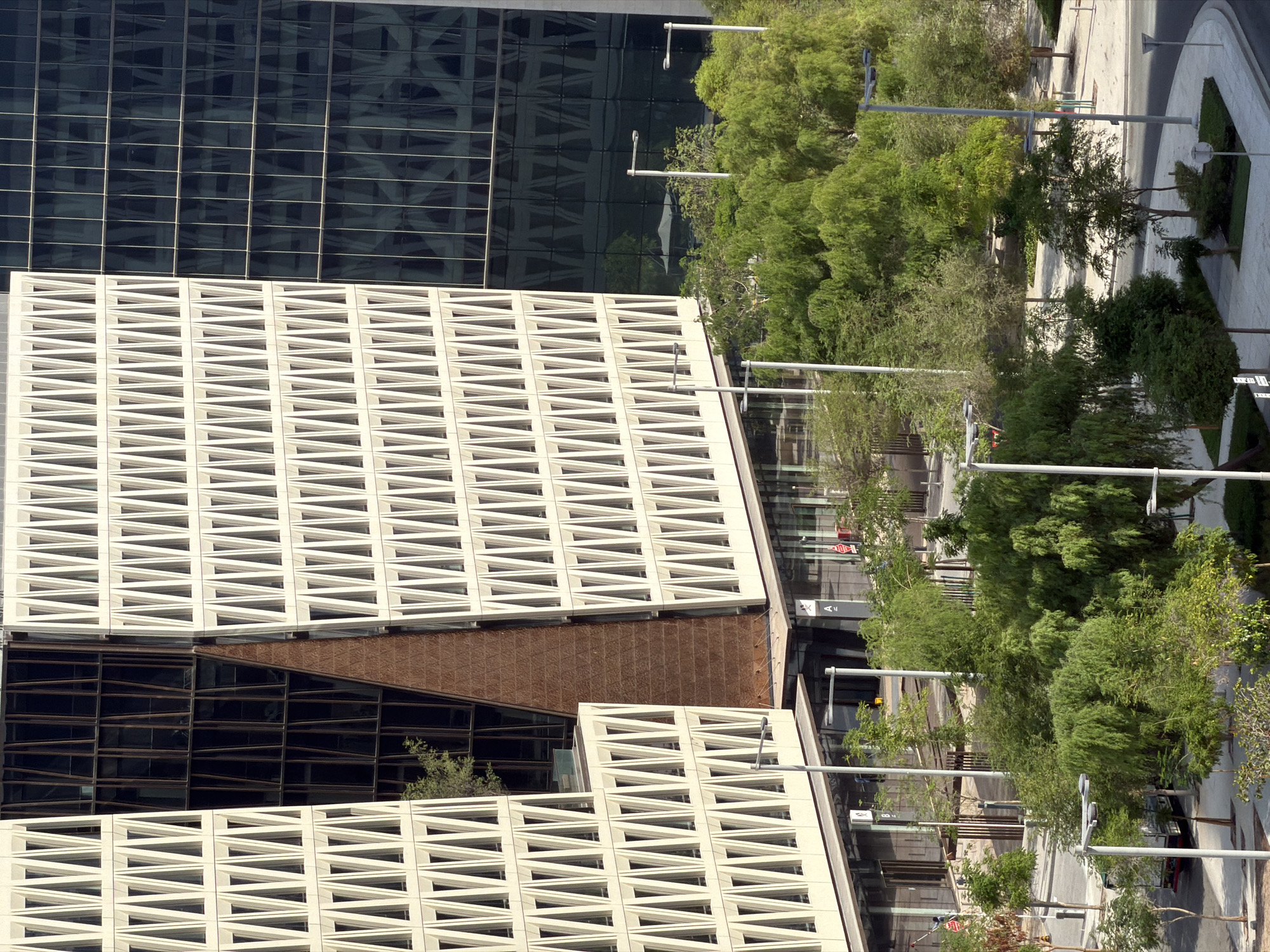 The image shows the exterior of a modern office building with a distinctive facade. The building features a repetitive pattern of triangular openings on its upper levels, contrasting with a darker, glass-fronted section on the right. The lower levels of the building appear to have commercial storefronts. Surrounding the building is a landscaped area with numerous trees and greenery, with paved walkways and streetlights visible. A bus is parked on the street in the lower left portion of the image.