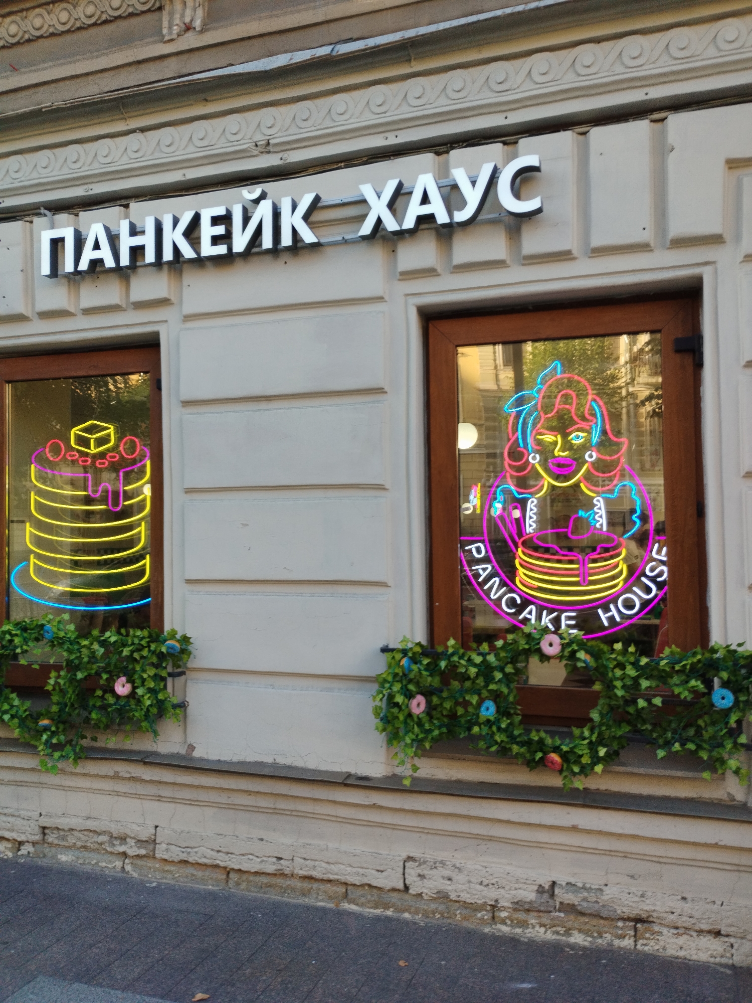 The image shows a storefront in Saint Petersburg, Russia, with two large windows and a prominent sign. The sign, in white block letters with a black outline, reads "ПАНКЕЙК ХАУС" (Pancake House).
The left window displays a neon sign shaped like a stack of pancakes with a pat of butter on top, dripping syrup, and garnished with cherries. The neon colors are yellow for the pancakes and butter, pink for the syrup and cherries, and blue for the plate.
The right window features a larger, more elaborate neon sign of a woman with vibrant pink and cyan hair, wearing a dress, winking, and holding a pancake. This sign also encircles a stack of pancakes and reads "PANCAKE HOUSE" in pink neon lettering.
Below both windows, there are window boxes filled with artificial green ivy and decorated with colorful donut-shaped ornaments in pink, blue, and yellow. The building facade is made of light-colored, textured stone with decorative moldings. The sidewalk in front of the building is paved with grey asphalt, and a few fallen leaves are visible. The overall scene suggests a bright and cheerful establishment.