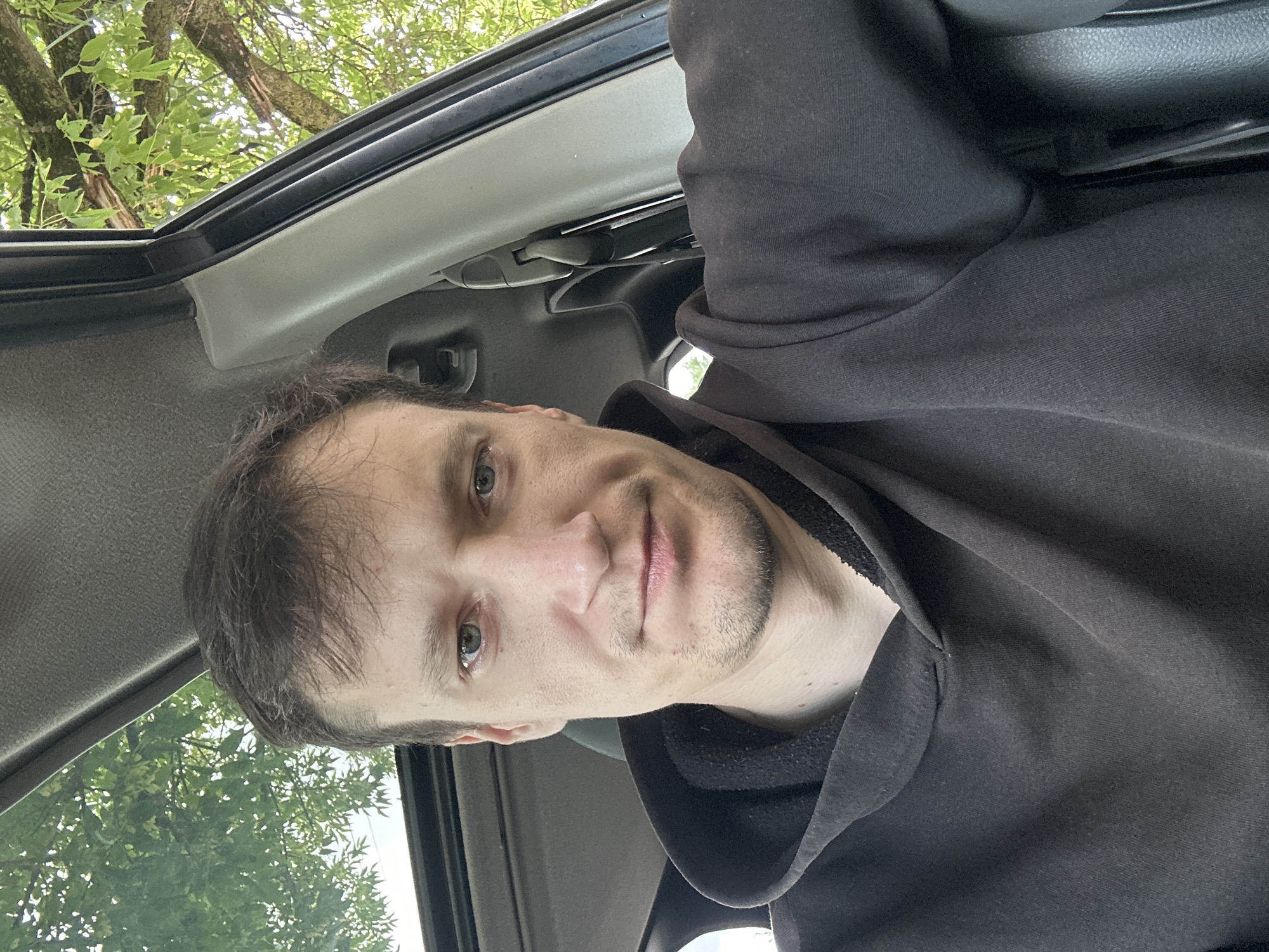 A close-up, head-and-shoulders selfie taken from the inside of a vehicle shows a man with short, brown hair and light green eyes. He is wearing a dark grey hooded sweatshirt. The man is looking directly at the camera with a neutral expression. Through the car's sunroof and open window, green foliage from trees is visible against a bright sky, suggesting an outdoor setting, possibly in the city of Makiv, Ukraine. The interior of the car is dark, with the grey headliner and a black seat belt buckle visible.