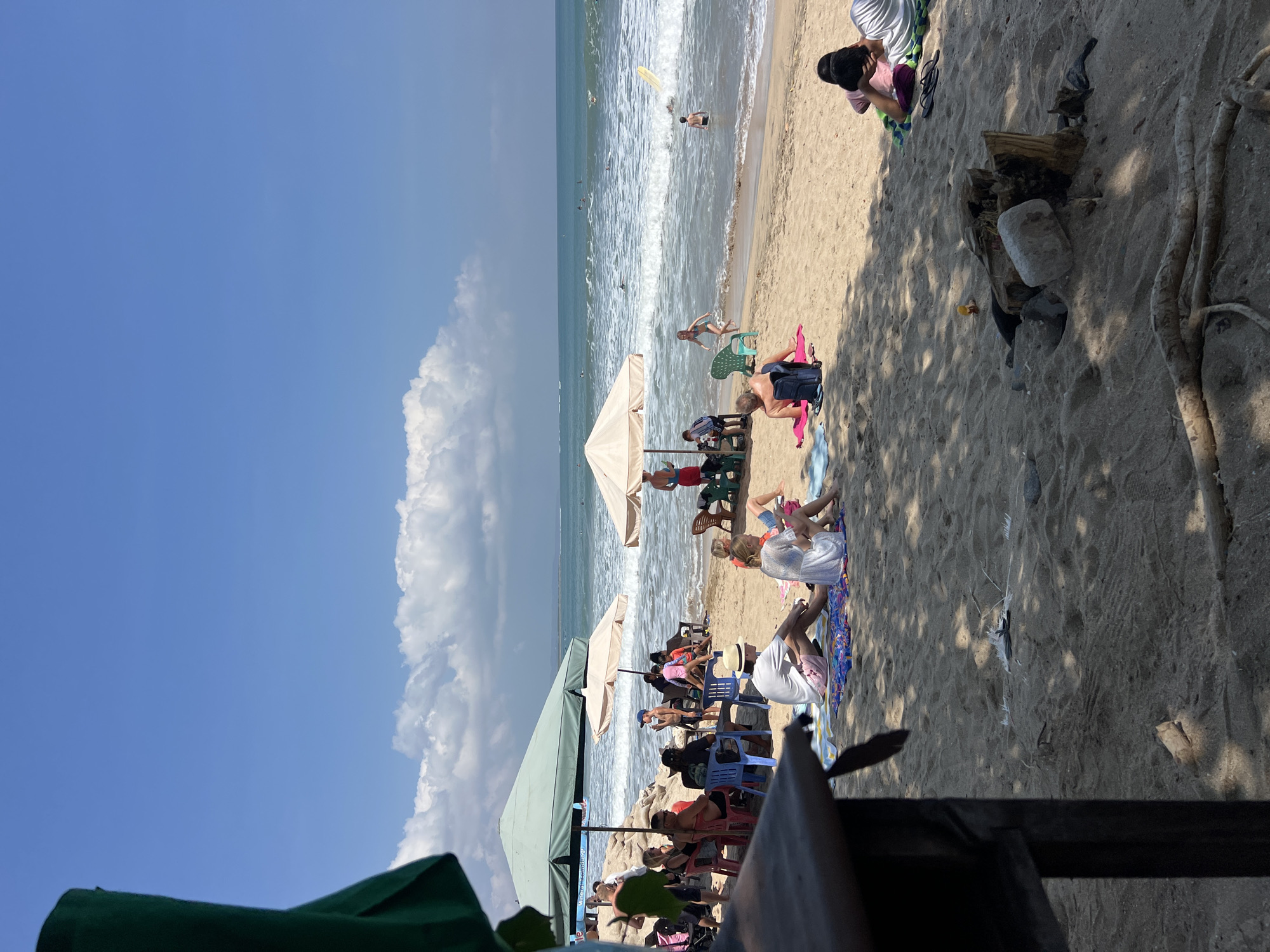 The image captures a sunny day at Kuta Beach, Indonesia. In the foreground, a dark wooden structure, possibly a beachside stall or table, casts a shadow over the sandy ground. Scattered on the sand are pieces of driftwood and some debris, including a dead bird.
Further back, numerous people are enjoying the beach. Several individuals are seated under large umbrellas, either on chairs or directly on the sand. Some are engaged in conversation, while others appear to be relaxing. A few people are actively involved in water activities, with surfers visible in the ocean, riding waves. Children are also present, with one girl running towards the water.
The sea is a prominent feature, with visible waves and a clear blue sky above. The horizon line shows a faint landmass in the distance. The overall scene depicts a typical, lively beach atmosphere in Kuta.