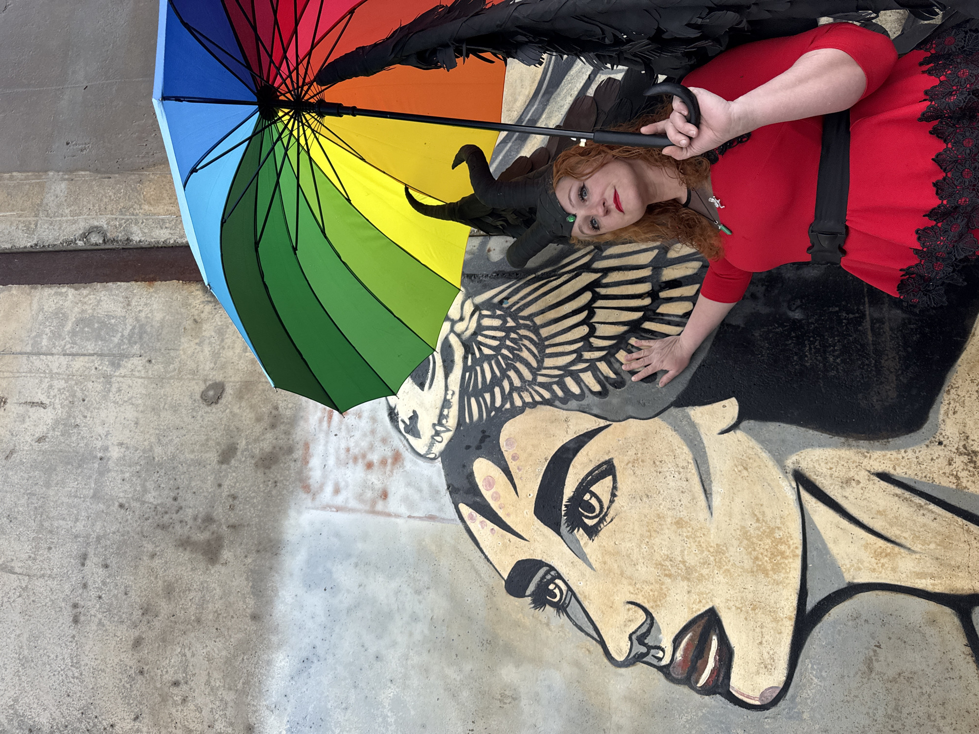 A red-haired woman in a red dress with black lace trim, a black belt, and a black horn headpiece and black wings lies on a concrete surface with a mural of a stylized woman's face and wings. The woman holds a rainbow-colored umbrella over her head. The mural appears to be painted on a concrete floor or surface. The location is likely Chaikovsky, Russia, based on prompt information.