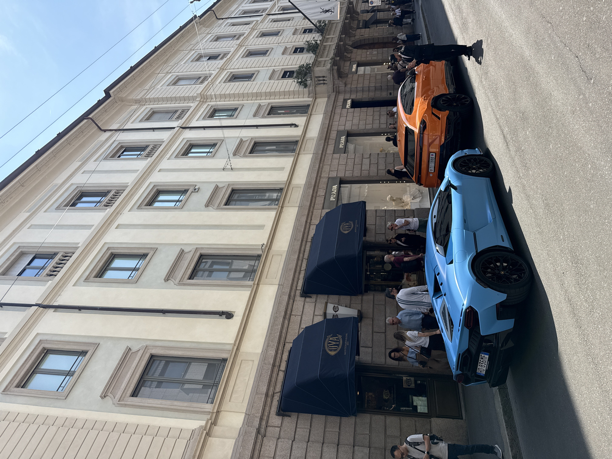 The image captures a street scene in Milan, Italy. A light-blue Lamborghini and an orange SUV-style vehicle are parked along the roadside. A building with multiple windows and stone trim lines the street, with "PRADA" signage visible at street level. People are visible along the sidewalk in front of the building. An awning with "OYVA MONTE NAPOLEONE" is visible above a doorway. A man is seen near the light-blue Lamborghini.