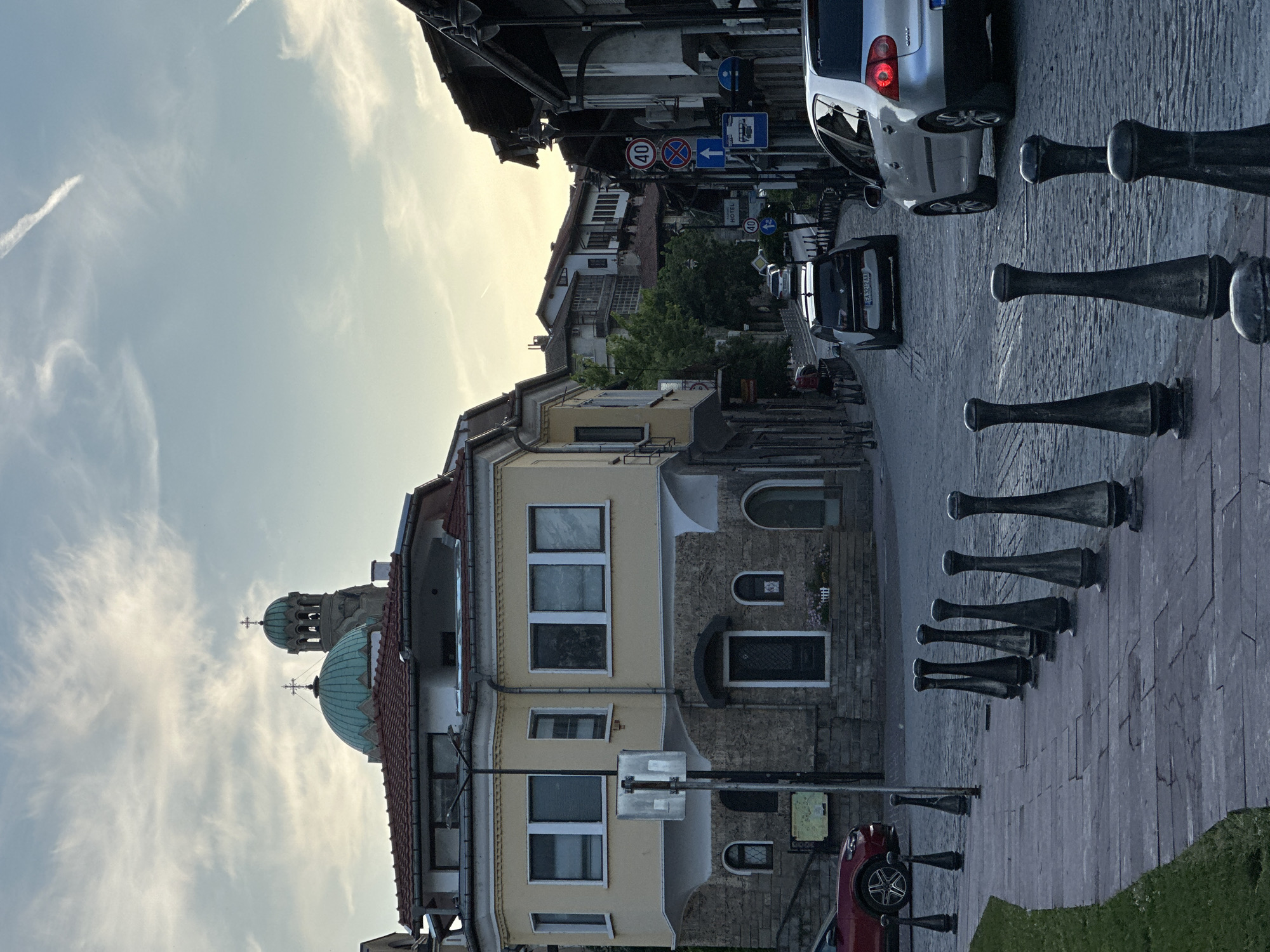 The image shows a street scene in Veliko Tarnovo, Bulgaria. In the foreground, a cobblestone street is lined with decorative bollards. A red car is parked on the left, and a silver car is driving on the right. In the background, a yellow building with a red-tiled roof is visible, along with a church with a green dome and bell tower. The sky is cloudy with some wispy clouds. The overall impression is of an old European city with a mix of residential and religious architecture.