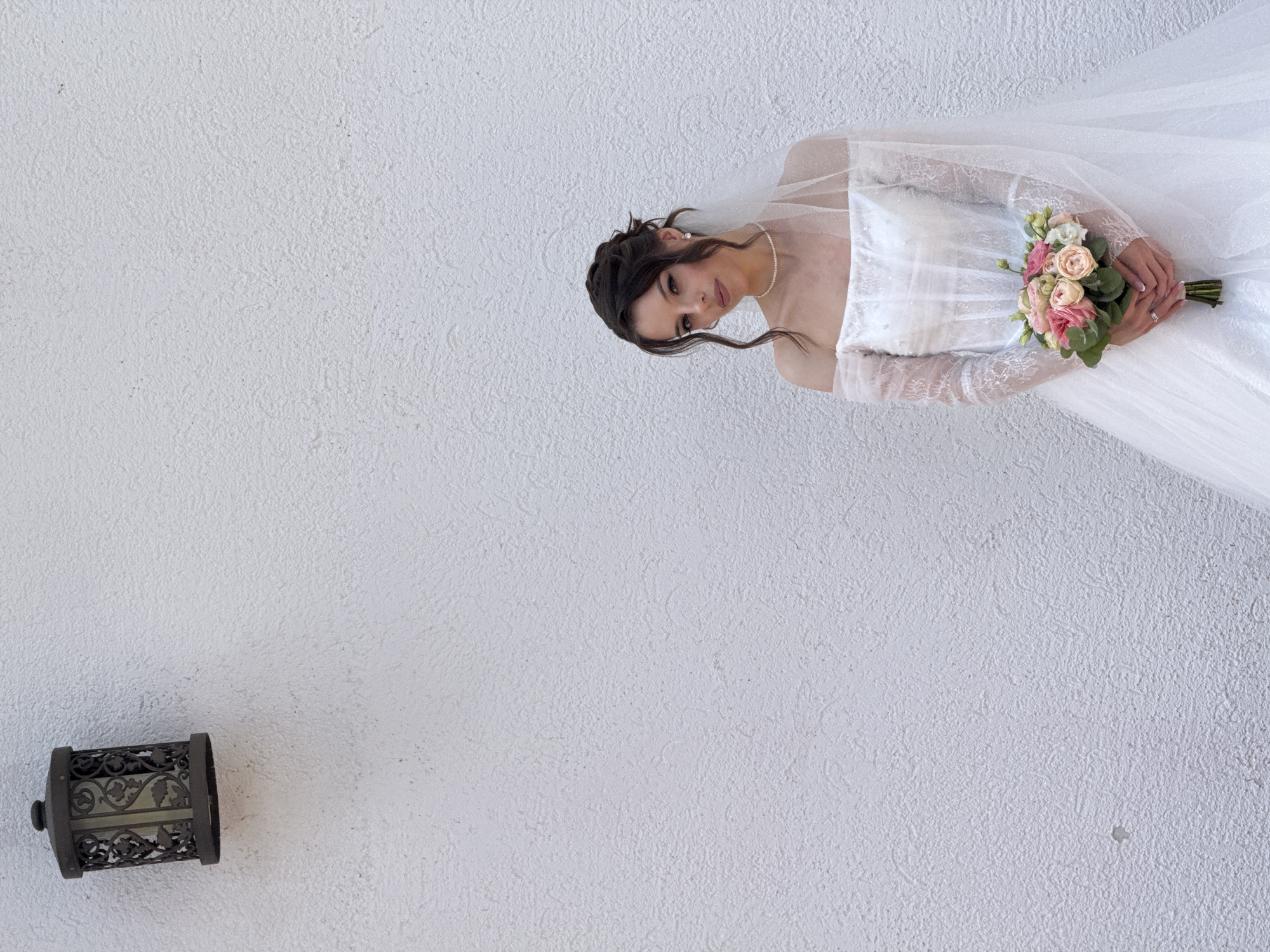 A young woman, dressed in a white wedding gown with off-the-shoulder lace sleeves and a veil, holds a bouquet of pink and white roses. She is standing against a textured white wall, with a vintage-style lantern mounted on the wall to her left. The woman has dark hair styled in an updo with loose tendrils framing her face. She is wearing a pearl necklace and earrings. The scene is indoors, likely a studio or a venue with a plain white backdrop. There is no indication of the city of Votkinsk, Russia, or any specific urban environment.