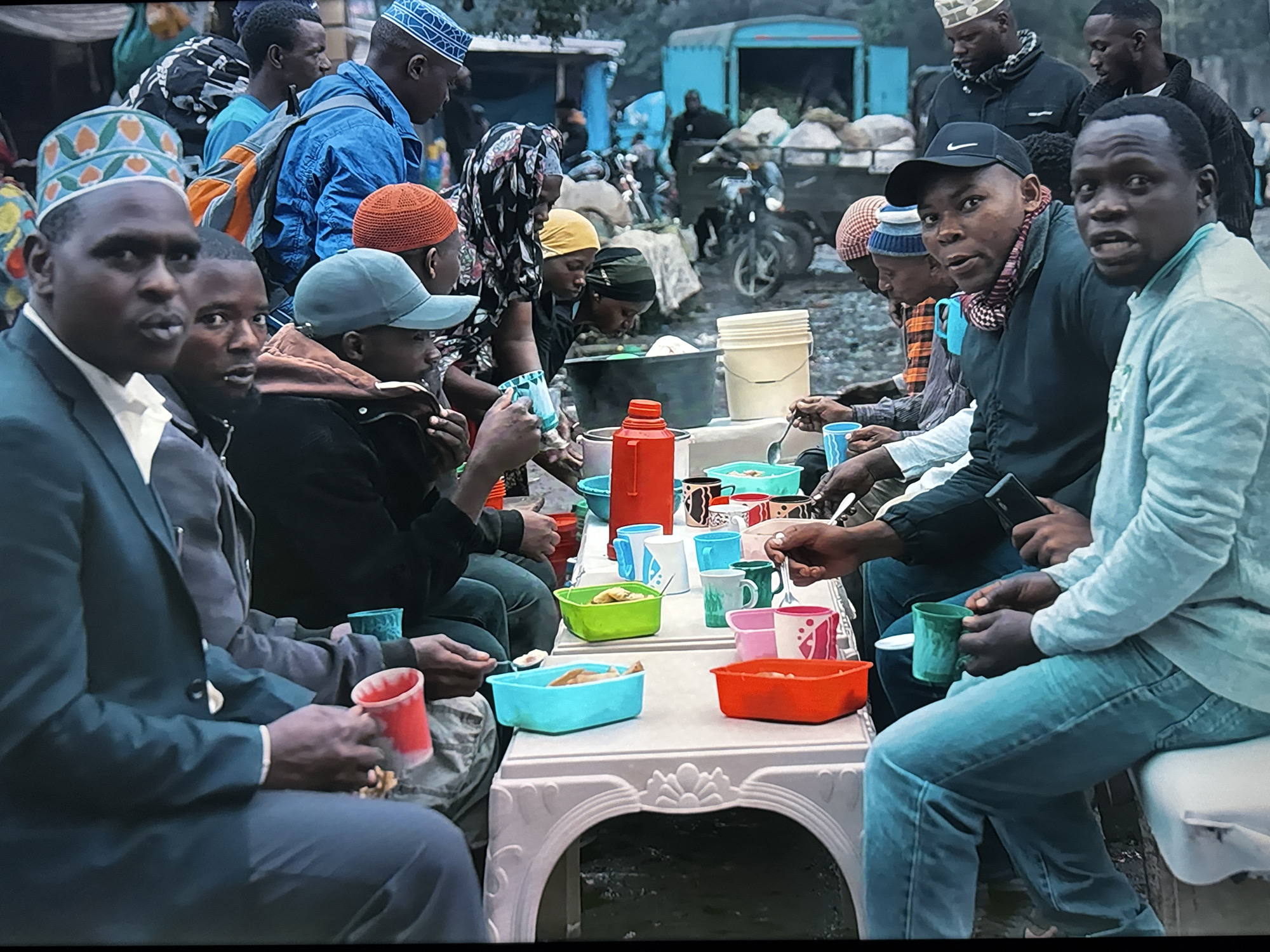 The image depicts a group of men and women gathered around a low table, possibly in a market or an outdoor gathering area. The individuals are of African descent, dressed in casual clothing, and some are wearing traditional head coverings. On the table, there are various bowls and cups containing food and drinks, indicating a meal or snack time. Several individuals are holding utensils and appear to be eating or serving themselves. In the background, there are elements suggestive of a bustling environment, including what looks like a truck, motorcycles, and other people, possibly indicating an urban setting. The overall atmosphere appears to be informal and communal.