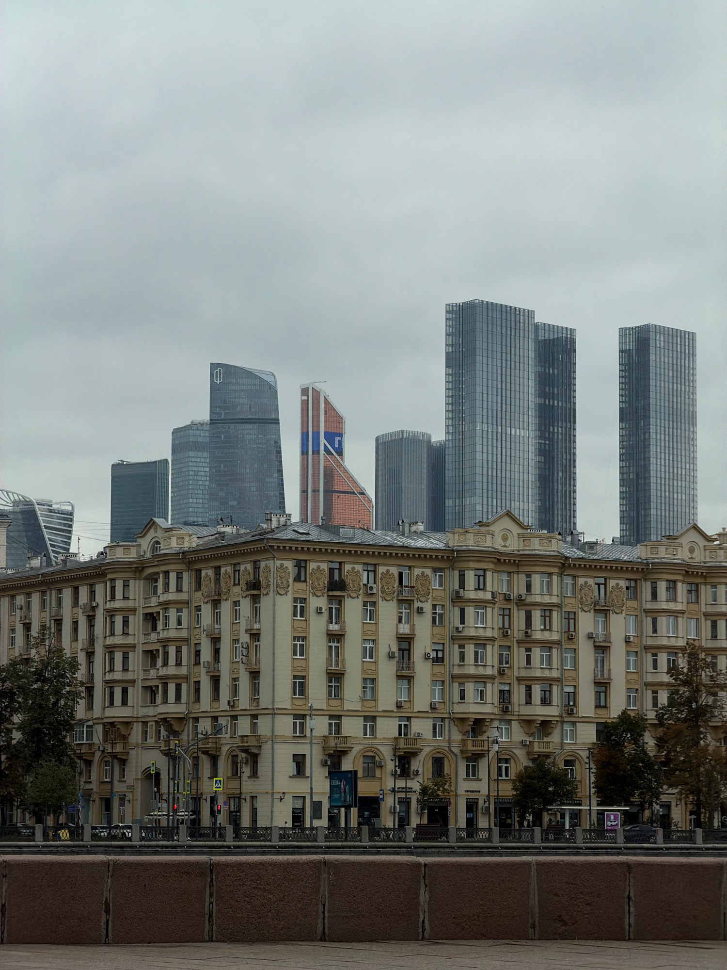 The image depicts a street-level view of the Moscow International Business Center, also known as Moscow-City, an area of skyscrapers in Moscow, Russia. In the foreground is a historic, multi-story apartment building with cream-colored facade, adorned with decorative architectural elements. Behind this building, several modern skyscrapers rise into a cloudy, overcast sky. Notable among the skyscrapers are the Neva Towers, the Federation Tower, and the Meriton-Nikolskaya Tower. The scene is bordered by a stone embankment in the lower part of the image, suggesting the presence of a river or canal. A street with traffic lights and some pedestrian activity is visible in the middle ground. The overall atmosphere is somber due to the overcast weather.