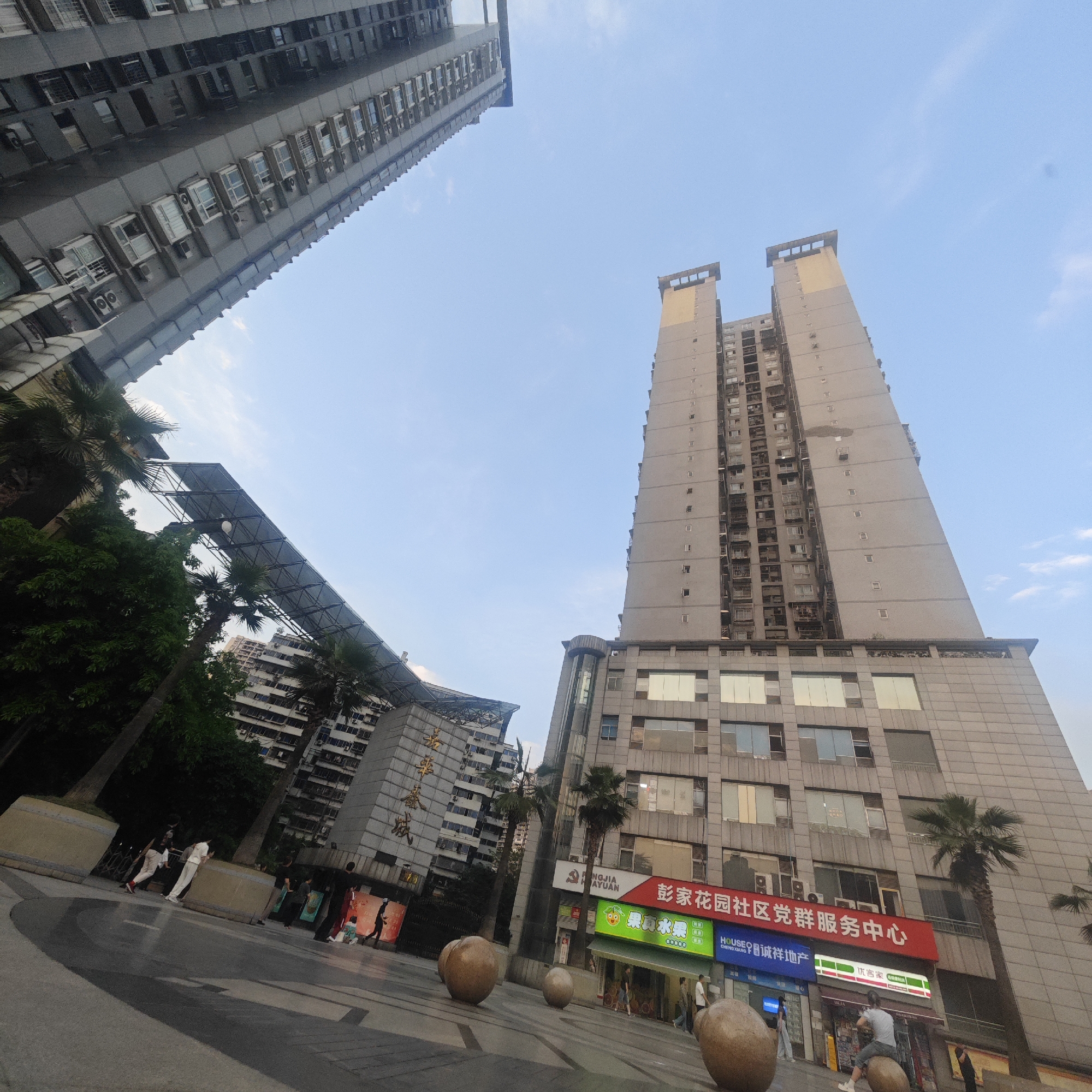 A low-angle wide shot captures a cityscape in Yuzhong District, China, revealing two tall, gray-toned buildings against a backdrop of a clear, light blue sky. The building on the left is more densely packed with smaller windows and air conditioning units, viewed at an angle that suggests it is either extremely tall or positioned at a slant relative to the camera. To the right, the second building is taller and straighter, with a tiered design. Its lower stories feature a mix of glass and solid panels, transitioning upwards into a series of vertically stacked apartments or offices.
At ground level, the shot captures a public space adorned with palm trees and geometric shade structures. People are seen walking and interacting in this space, creating a sense of activity and daily life. The foreground features several rounded, brown sculptures scattered across the plaza, adding an artistic touch to the urban landscape. In addition, stores and businesses are situated in the buildings next to the plaza. Specifically, the storefronts are decorated with Chinese signage, which includes the names of stores, the name of the district (Pengjia Garden), a political slogan, and a real estate company.