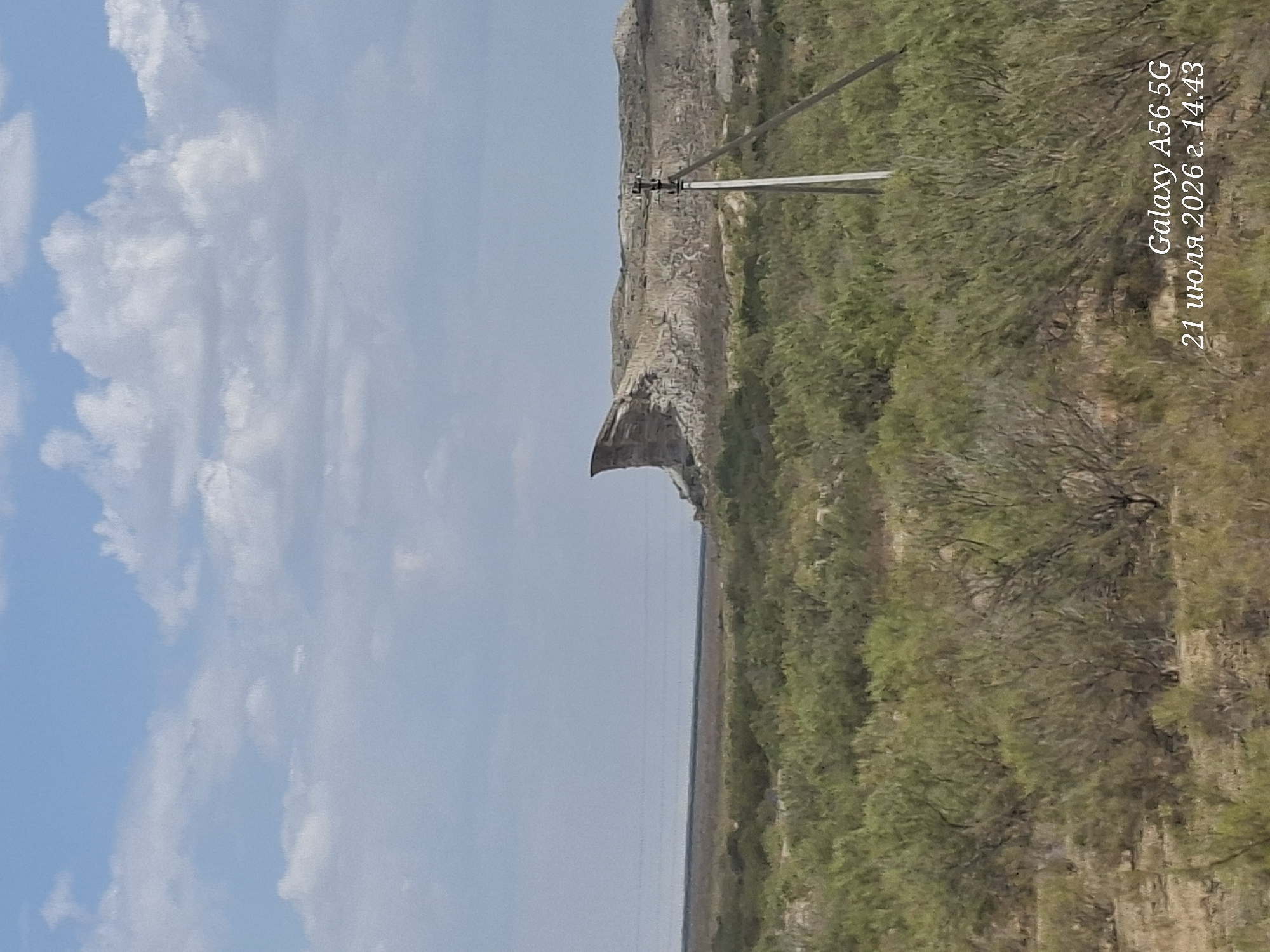 The image depicts a vast, arid landscape under a partly cloudy sky. The foreground is dominated by sparse, green shrubs and dry, brownish ground. In the midground, a large, weathered rock formation with distinct layered strata rises prominently. To the right of the rock formation, a solitary utility pole with wires is visible. The background features a hazy horizon with a flat, treeless plain. The bottom right corner of the image contains a timestamp and device model, "Galaxy A56 5G 21 июля 2026 г. 14:43," which translates to "July 21, 2026, 14:43." The text suggests the location is in Elabad, Uzbekistan, a city known for its desert environment and geological formations.