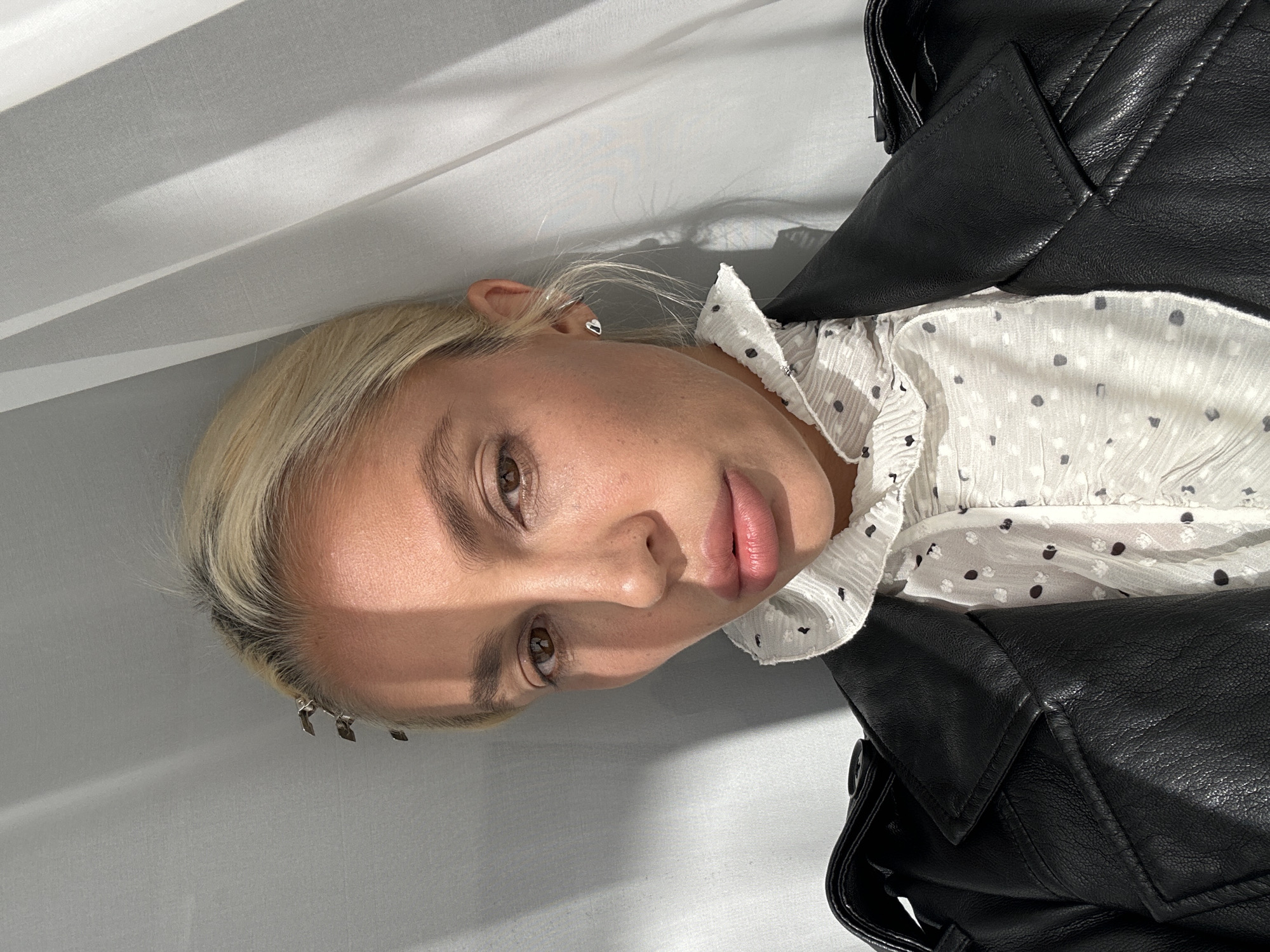 A woman with blonde hair and light skin is looking towards the camera. She is wearing a white ruffled shirt with a polka dot pattern and a black leather jacket. Her hair is pulled back and she has a hair clip on the side of her head. She is also wearing a heart-shaped earring in her visible ear. The background is a light-colored curtain with shadows cast on it. The lighting suggests it is daytime. The location of Kuta Selatan, Indonesia is not depicted in this image.
