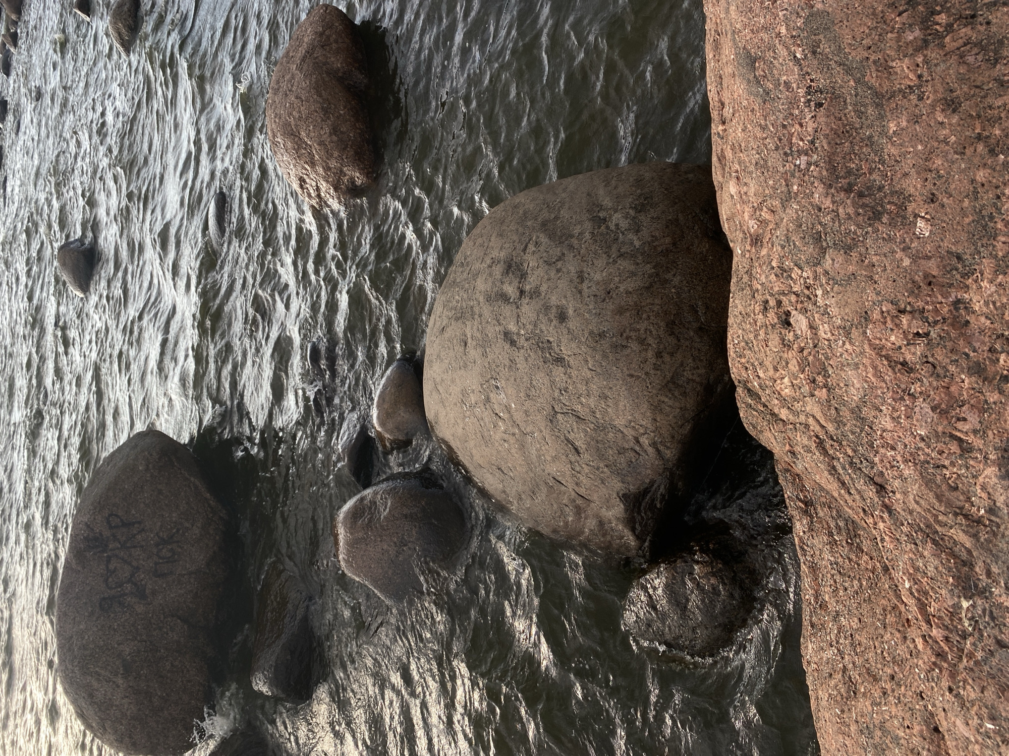 In the city of Baltiiskoe, Russia, a body of water, likely the Baltic Sea, is visible with choppy waves. Several large, round boulders are partially submerged in the water, with their tops exposed. One large boulder in the foreground has graffiti on its surface, including what appears to be letters and a heart symbol. In the lower right corner of the image, a large, rough-textured reddish-pink boulder is prominent, with a portion of a larger rock formation visible behind it. The lighting suggests it might be late afternoon or early evening, with a warm glow reflecting on the water and rocks.