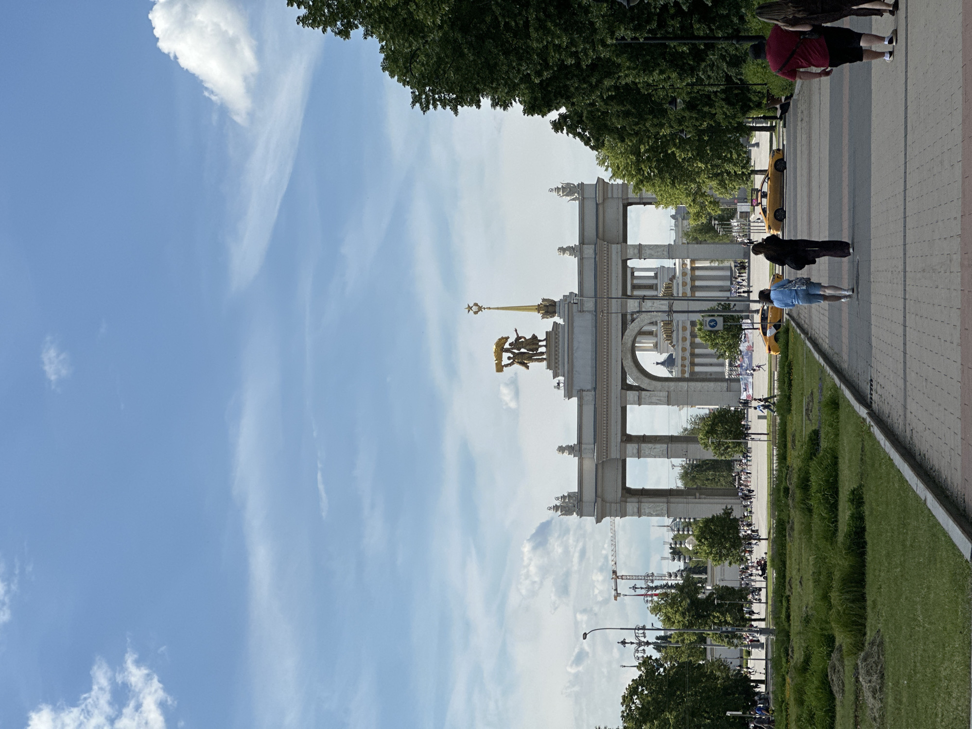 The image presents a grand outdoor scene under a bright, sunny sky with scattered white clouds. The primary subject is a majestic, ornate architectural structure, resembling a triumphal arch, positioned centrally in the background. This elaborate entrance, characteristic of the Main Entrance Arch of VDNKh in Moscow, Russia, features multiple large archways and classical detailing.
Crowning the structure is a prominent golden sculptural group, depicting figures (likely workers or collective farmers), beneath a tall golden spire topped with a five-pointed star, all glinting in the daylight. The light-colored stone of the architecture contrasts with the clear blue sky.
In the mid-ground and foreground, a wide, paved walkway stretches towards the arch, bordered by vibrant green grassy areas and mature trees with lush foliage. Several people are visible on the walkway, engaged in casual strolls. Notable individuals include a person in a red shirt and black shorts, another in a blue top and light shorts, and a third in a dark coat. Two yellow vehicles, possibly small electric shuttle carts, are seen on or near the paved area, one closer to the arch and one further down the path. Decorative streetlights are also visible along the walkway.
Through the arches in the distance, other buildings and faint outlines of what appear to be construction cranes can be seen, indicating the vastness of the area beyond the entrance. The time of day is clearly daytime, with strong natural light suggesting late morning or afternoon. The weather is clear and pleasant. There is no discernible text visible in the image. The scene conveys a sense of a busy, yet relaxed, public park or exhibition ground.