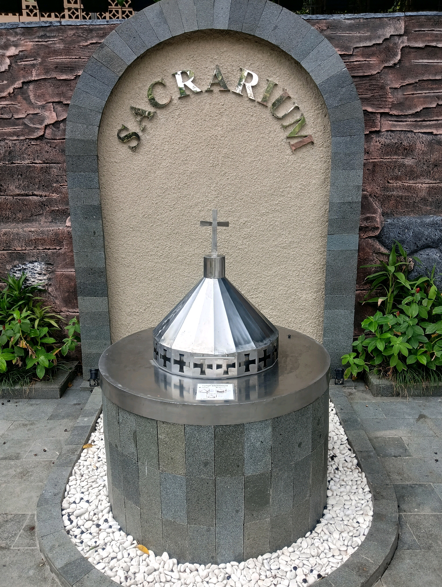 The image depicts a sacristy structure located in the city of Narimbang Mulia, Indonesia. The sacristy is a circular, stone-clad pedestal topped with a conical, stainless steel structure that culminates in a cross. The conical structure features decorative cross-shaped cutouts around its base. The entire monument is surrounded by white pebbles. Behind the sacristy, a textured, reddish-brown wall forms the backdrop, framed by a gray arched stone structure. The area is further enhanced by lush green plants flanking the sacristy on either side.
