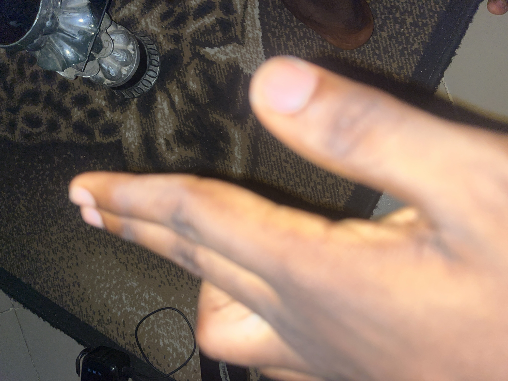 The image shows a close-up of a person's hand with fingers extended towards the camera, obscuring much of the background. The hand is positioned in the foreground, with a portion of what appears to be a foot or leg visible in the lower right corner. The background consists of a carpet with a pattern of brown, black, and beige. A silver decorative object, possibly a candle holder or vase, is visible on the carpet. To the left, a black electronic device with a coiled wire is partially visible, suggesting it might be a camera or other accessory. The lighting in the image is warm, casting a golden hue on the hand. The scene is domestic, likely within a home environment in Jalingo, Nigeria.