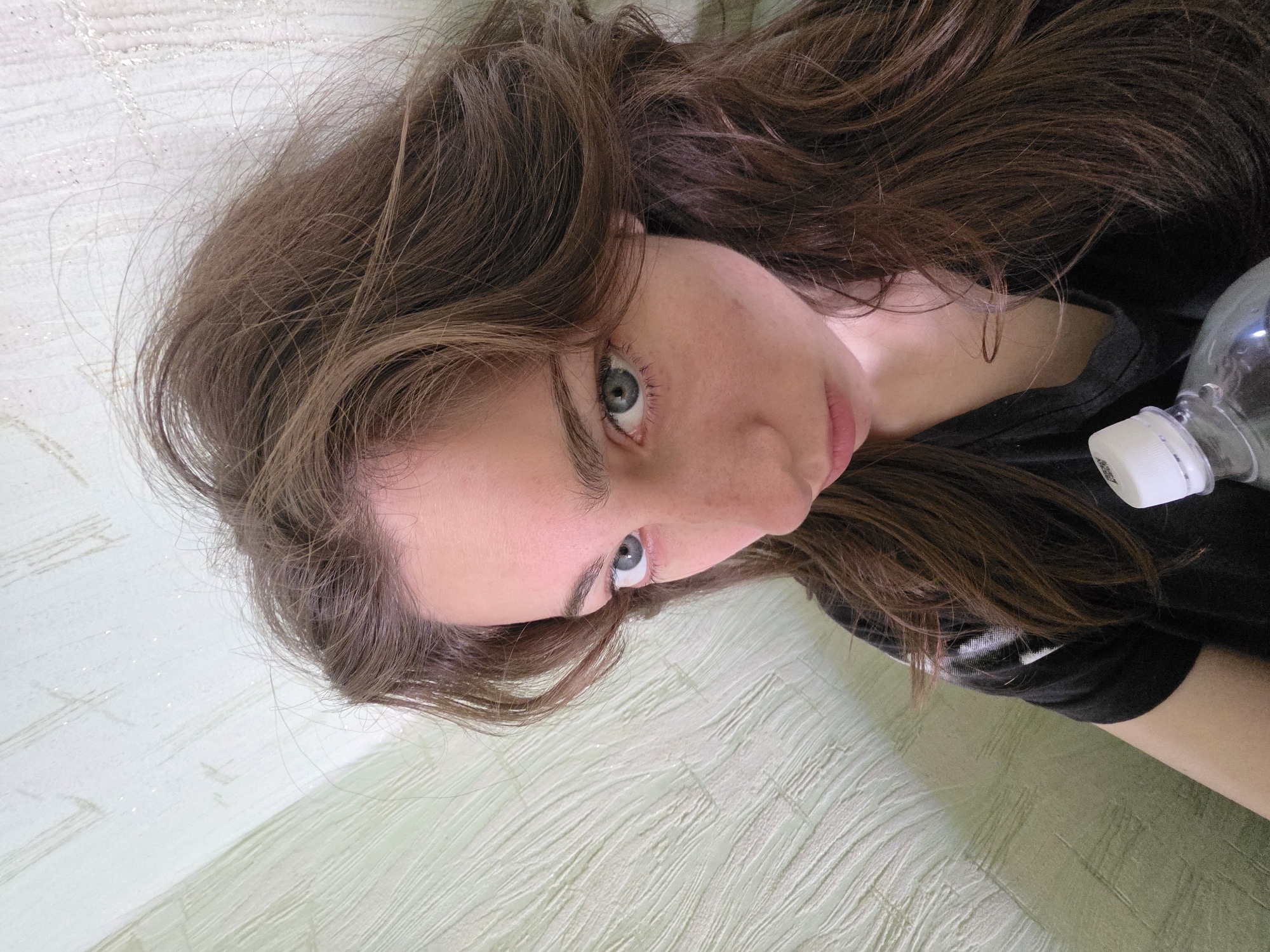 A young woman with long, wavy brown hair and blue eyes is captured in a close-up shot, looking downwards. She is wearing a black t-shirt. Her skin appears flushed. In the foreground, a clear plastic water bottle with a white cap is visible, held by the woman. The background consists of a textured, pale green wall. The overall lighting suggests an indoor environment, likely in an apartment. The image does not contain any information that would indicate the location is the city of Chita, Russia.