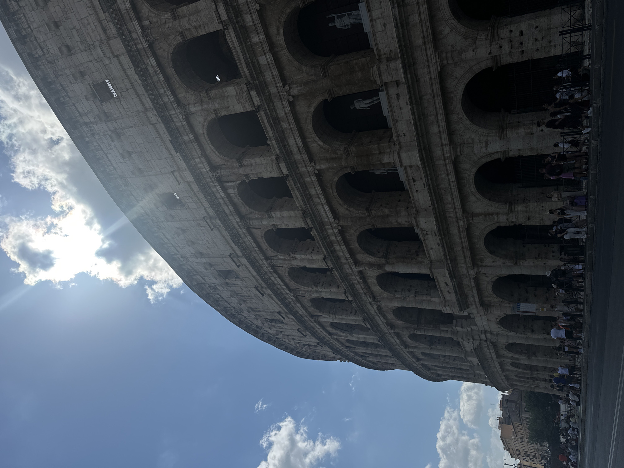 The image shows the Colosseum in Rome, Italy. The perspective is from a low angle, looking up at the ancient amphitheater on a partly cloudy day. The sun is visible in the upper left corner, with light rays piercing through the clouds. The Colosseum's stone facade, with its characteristic arches and tiers, dominates the right side of the frame. Some statues are visible within the arched openings on the lower levels.
In the foreground, a street runs along the base of the Colosseum, with a sidewalk and a fence separating it from the monument. A group of people, appearing to be tourists, are gathered on the sidewalk, some standing and others sitting. They are dressed in casual summer clothing. Behind the Colosseum and to the left, several buildings are visible, including a residential building with a light-colored facade and reddish-brown roofs. The overall scene depicts a popular tourist destination on a bright day.