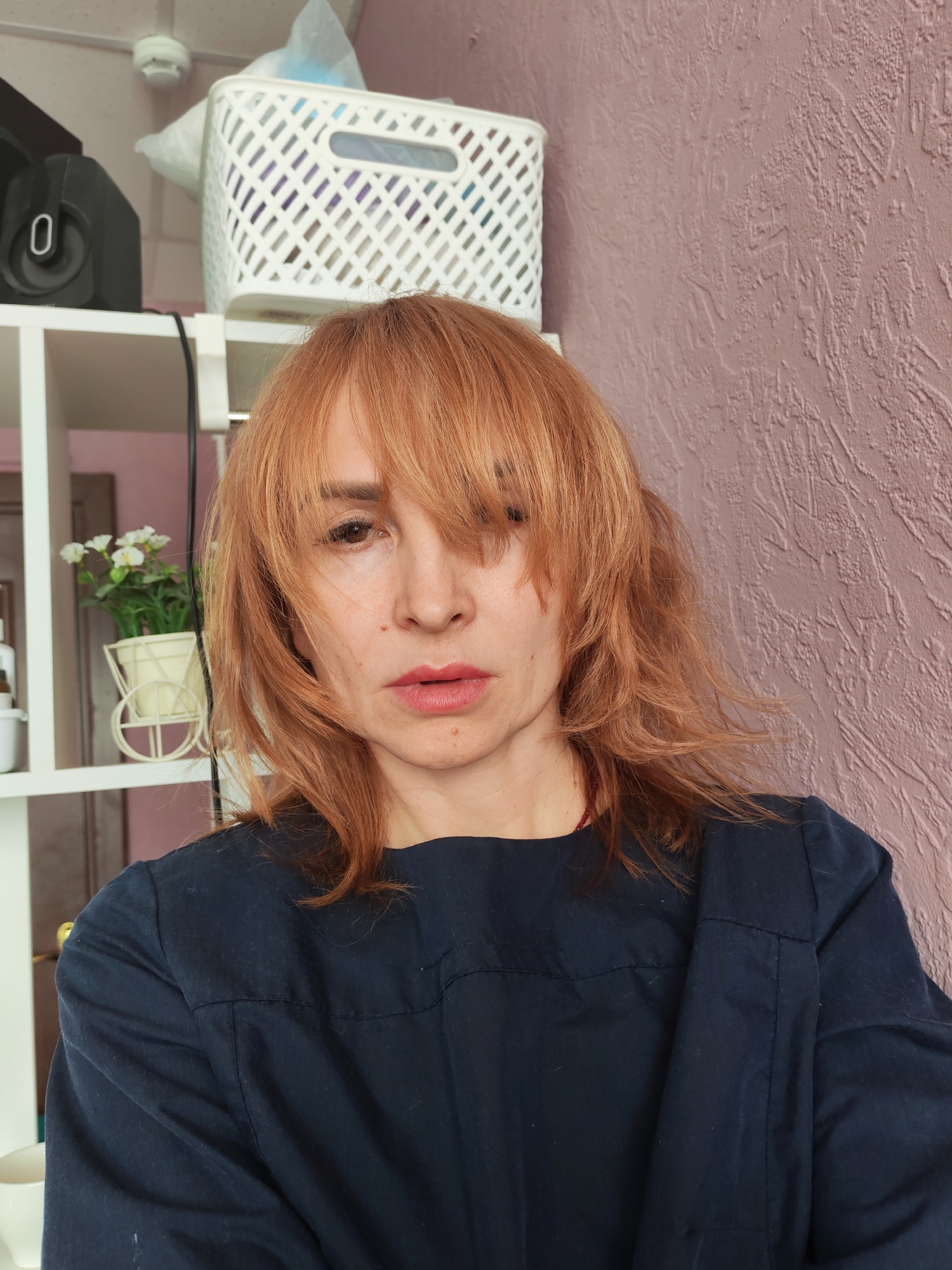 A woman with reddish-blonde hair stands indoors against a textured, mauve-colored wall. She wears a dark blue, long-sleeved shirt. Behind her, there's a white shelving unit with a black speaker, a white basket, and a small plant in a white cart-shaped planter. The location is indicated as the city of Chaikovsky, Russia.