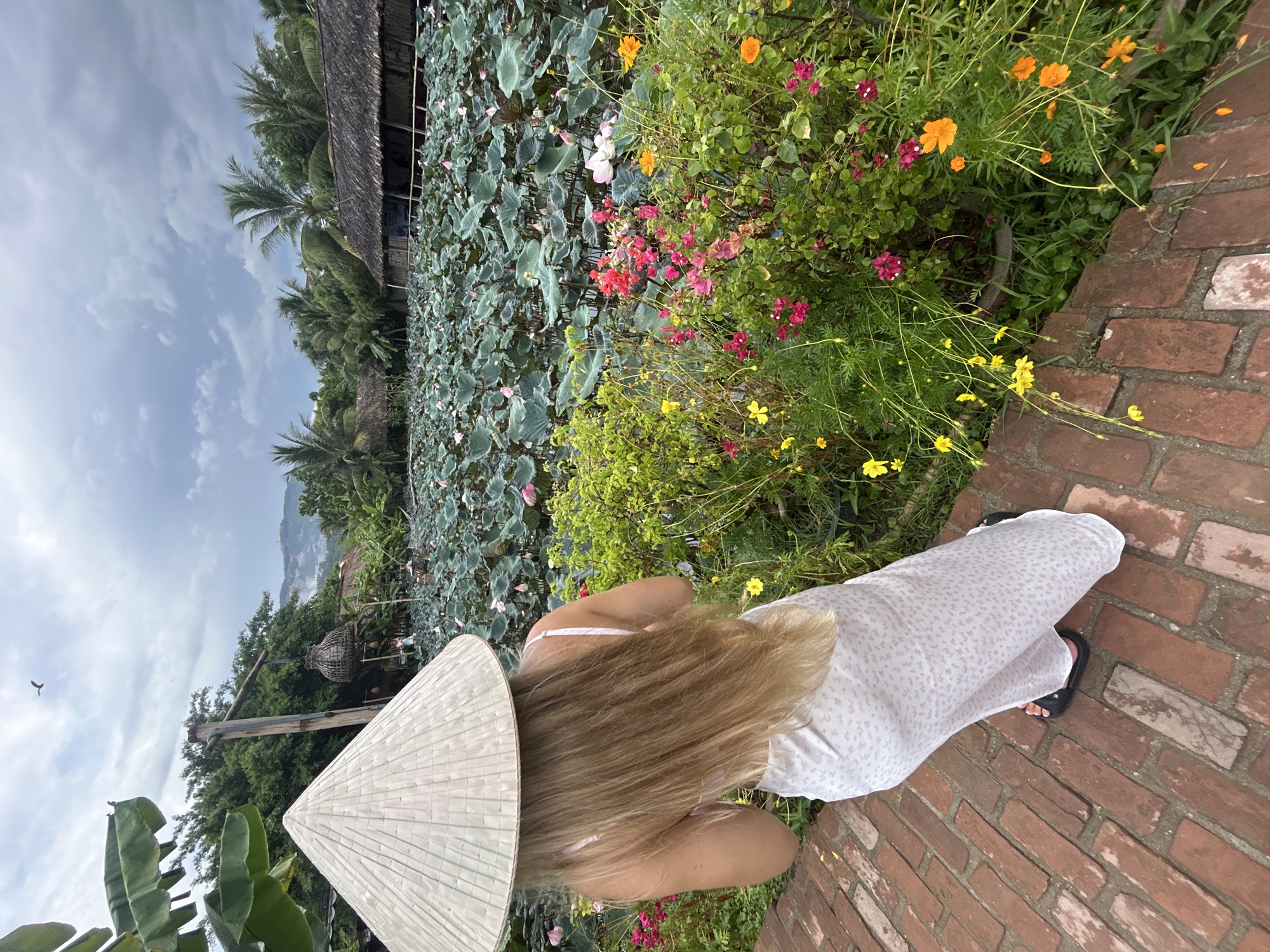 A woman wearing a conical hat and a white patterned dress stands on a brick pathway, facing away from the camera. Her long blonde hair is visible beneath the hat. In the background, a large pond is covered with lotus pads and some pink and white lotus flowers. To the right of the pond, there are lush green plants with small pink, red, yellow, and orange flowers. Beyond the pond, there are thatched-roof structures and palm trees, with hills visible in the distance under a cloudy sky. The scene appears to be a tranquil natural setting, possibly a garden or a rural landscape.