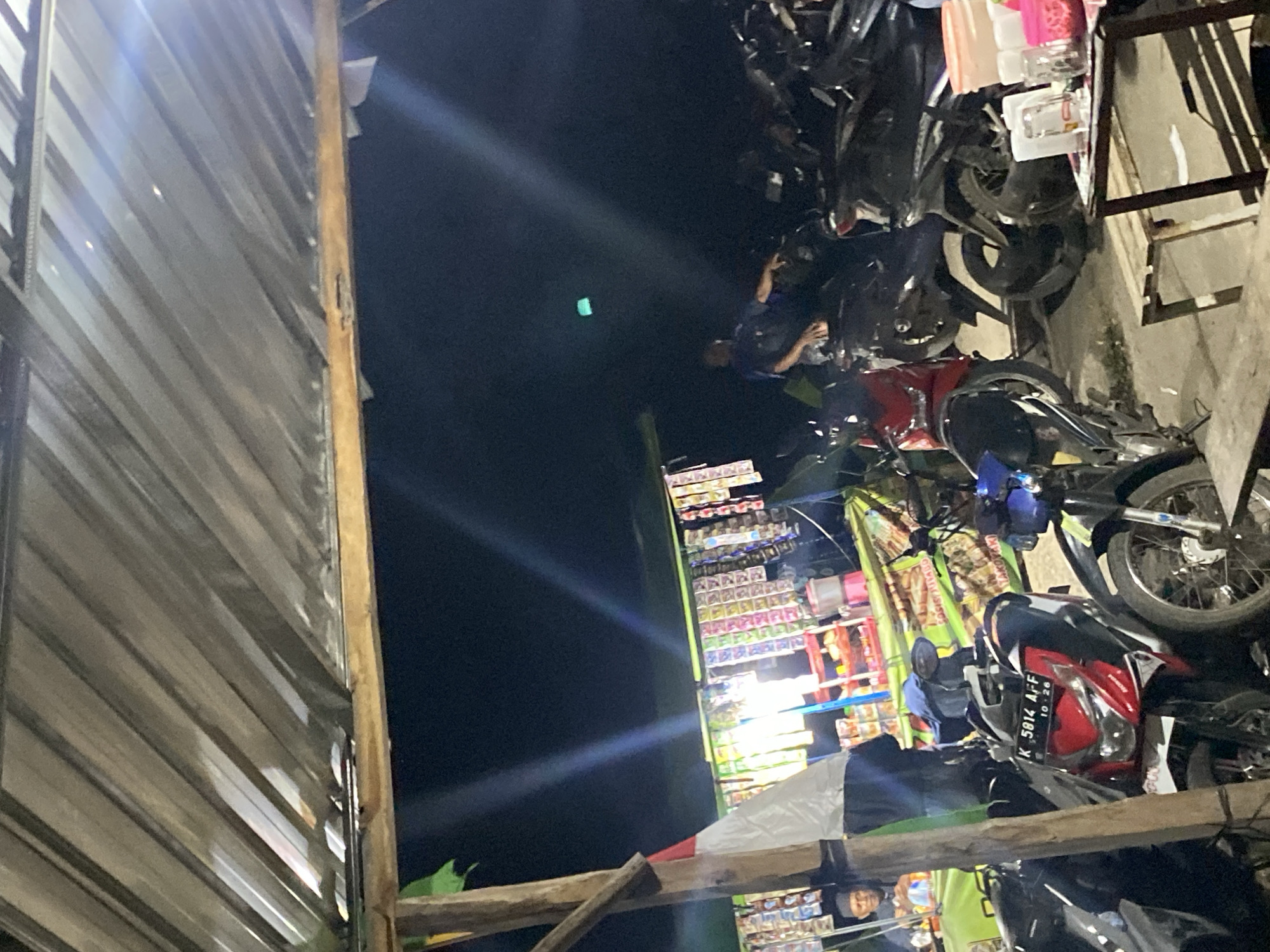 The image captures a nighttime scene, likely at a street-side stall or small marketplace in Unknown, Unknown. A corrugated metal roof, supported by wooden beams, covers part of the scene. Underneath, several motorcycles are parked, some in the foreground and others further back. To the left, a small stall is visible with racks of packaged goods, possibly snacks or cigarettes, hanging from the front. A man in a dark shirt and cap is seated at the stall. In the background, another man in a blue shirt is standing near a motorcycle. The lighting is dim, with bright streaks of light suggesting artificial illumination from the stall or other sources. A small table with cups and containers is set up in the lower right of the frame. The overall atmosphere appears casual and utilitarian, characteristic of an outdoor market or rest stop.