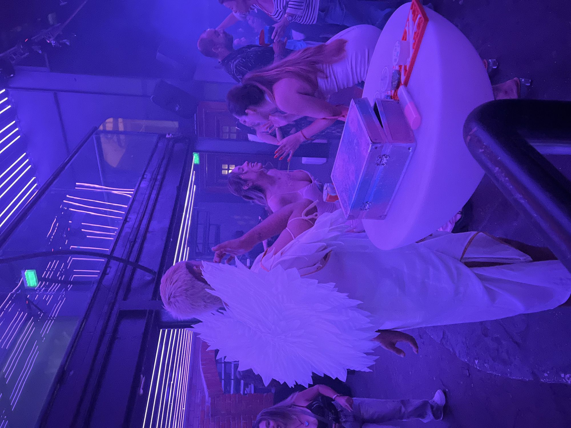 The image shows an interior scene, lit with blue and purple lighting, possibly a nightclub or event space, in Arica, Chile. A group of people are gathered around a round white table, on which there is a silver metal case and other objects. A person with white hair and a large white floral decoration stands at the edge of the table. Several women are interacting with the objects on the table, with their hands reaching toward them. One woman is seated on the floor near the bottom of the frame, and other people are visible in the background. The setting includes decorative lighting elements and structural features reminiscent of a building interior.