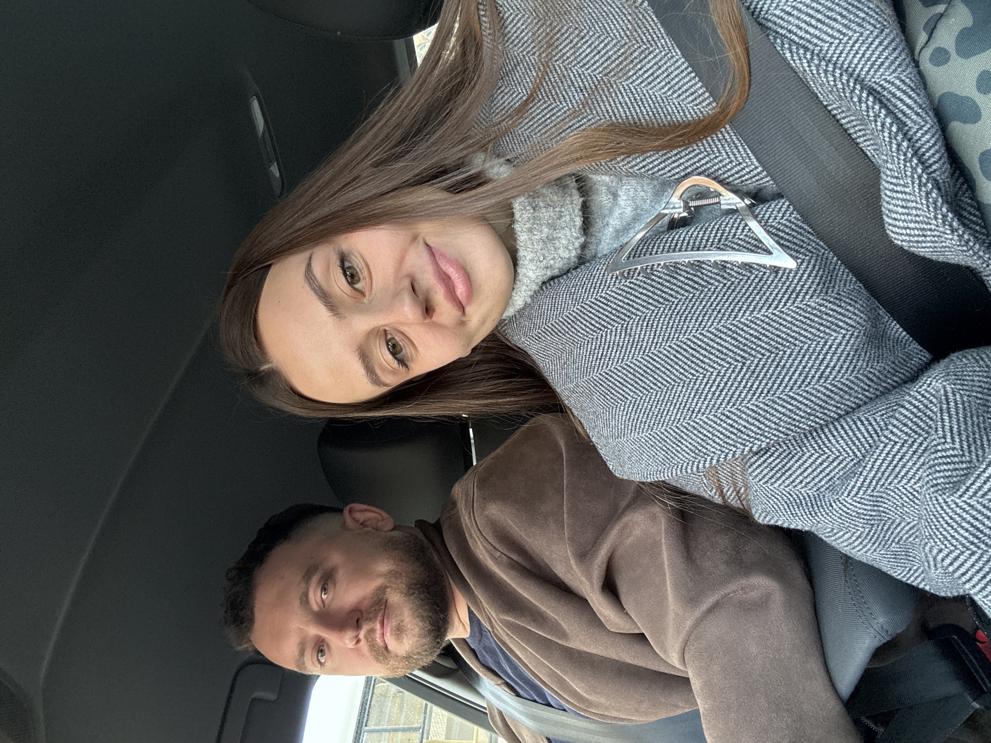 This tilted image captures a casual indoor portrait of two individuals, a woman and a man, seated within a vehicle, likely a car, in Krasnoyarsk, Russia.
In the foreground, a woman with long, straight brown hair, brown eyes, and fair skin looks directly at the camera with a gentle, slightly smiling expression. She is dressed in a grey ribbed turtleneck or sweater layered under a grey coat with a distinct herringbone pattern. A silver-toned, triangular hair clip is attached to her coat near her left shoulder. A dark car seatbelt is visible across her chest.
Behind and to her side, a man with short dark hair and a beard also looks towards the camera. He is wearing a brown coat over a dark blue garment. The car's dark interior, including the ceiling and parts of the seats, is visible around them. Through a window next to the man, a blurred outdoor scene with diffuse light, possibly indicating daytime, can be glimpsed.
The activity appears to be a casual selfie or photograph taken by the occupants. Both subjects convey a calm and relaxed mood. Their attire suggests cooler weather conditions. There is no visible text in the image.