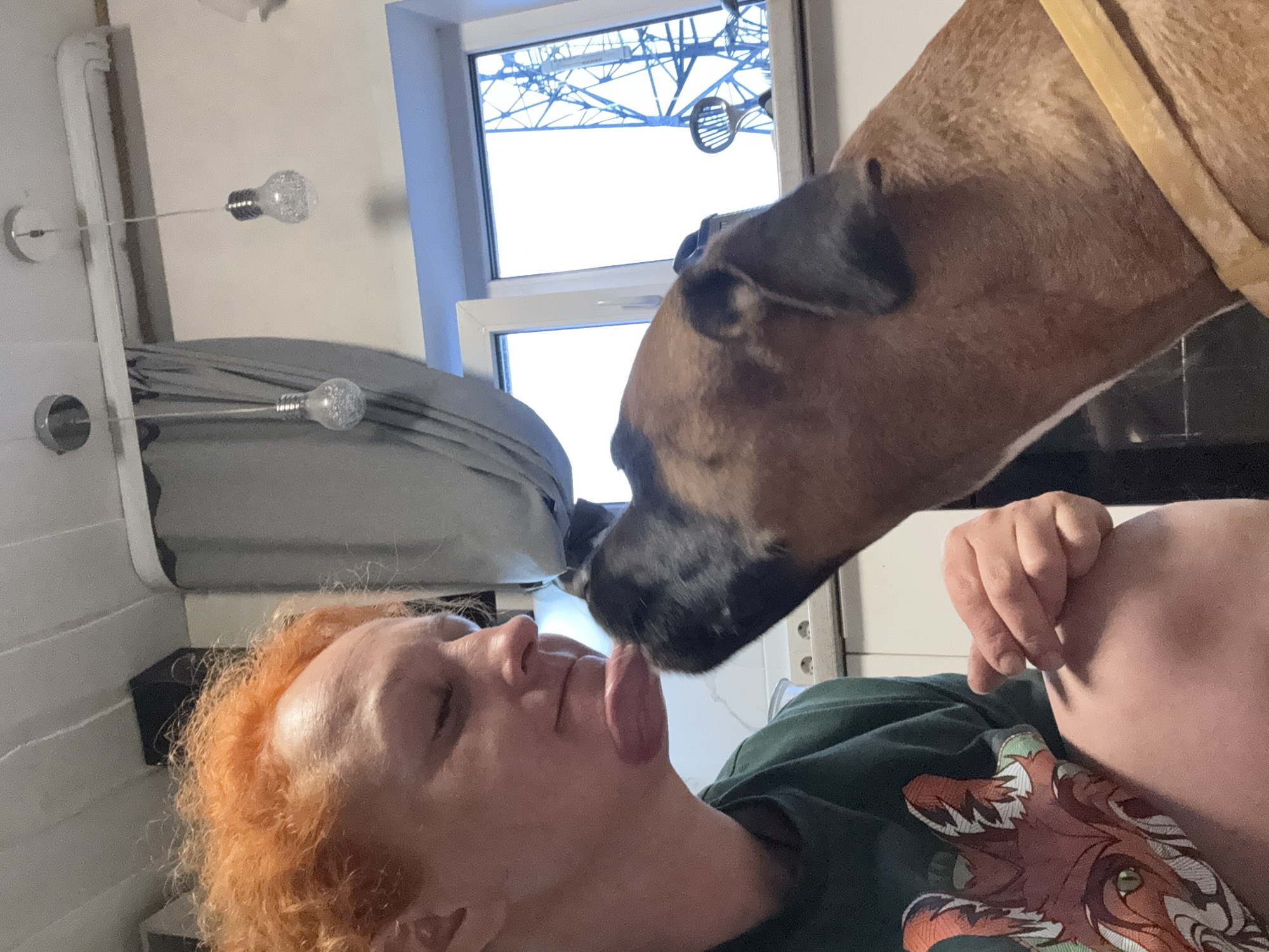 A photograph shows a woman with orange hair lying on her back while a brown dog licks her face. The dog is wearing a yellow collar. The woman is wearing a green shirt with an orange fox design. In the background is a window with a view of a metal structure. The photo was taken in Chaikovsky, Russia.