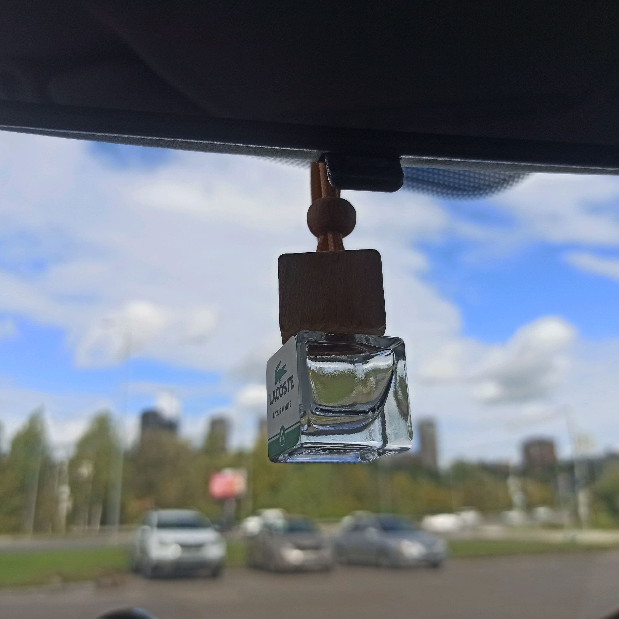 An air freshener shaped like a small glass bottle with a wooden cap hangs from the rearview mirror inside a car. The air freshener displays a label that reads "LACOSTE L.12.12 WHITE". Outside the car, the sky is blue with scattered white clouds. In the background, blurred buildings, trees, and several cars on a road are visible. The scene appears to be a daytime shot, possibly taken in an urban or suburban area. The image composition suggests the photo was taken by a passenger or the driver of the car, looking forward through the windshield.