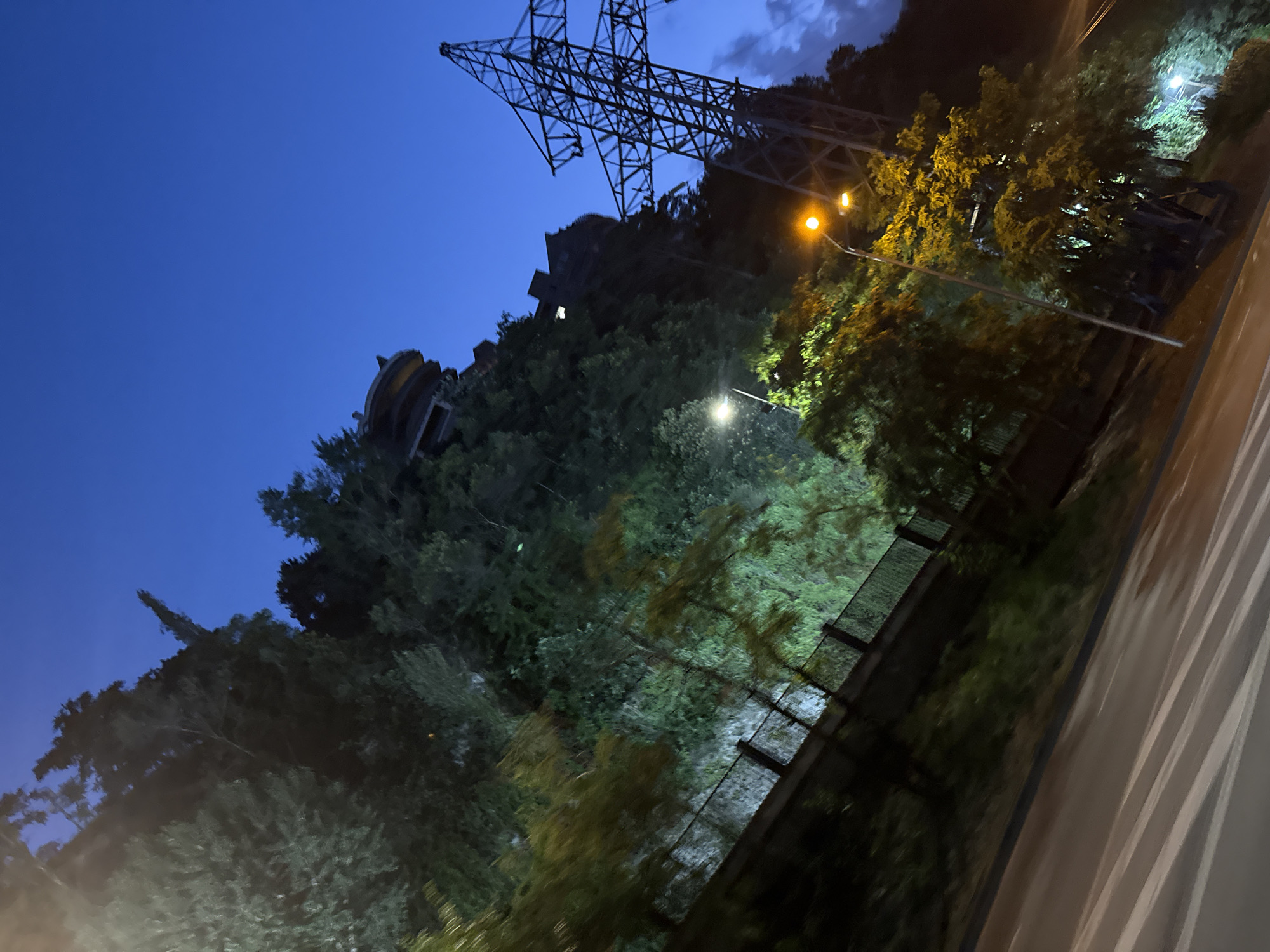 The image captures an evening scene in Tbilisi, Georgia, near a prominent transmission tower. The sky is a deep blue, indicative of twilight or early night. On the left, a steep, tree-covered embankment rises, topped by a round building with a distinctive architectural style, possibly an observation deck or part of a complex on the hill. A black metal fence runs along the base of the embankment.
The right side of the image is dominated by a tall, lattice-style transmission tower, a common fixture in urban and industrial areas. Its dark silhouette stands out against the twilight sky. Several streetlights are visible, casting pools of yellow and white light on the trees and road below. One streetlight, near the transmission tower, illuminates the foliage with a warm glow.
In the foreground, a section of a road is visible, with white pedestrian crossing lines blurred due to motion. The overall impression is a dynamic urban landscape at dusk, blending natural elements like trees and hills with imposing man-made structures. The tilt of the image suggests it was taken from a moving vehicle, adding to the sense of dynamism. No people are directly visible in the frame.