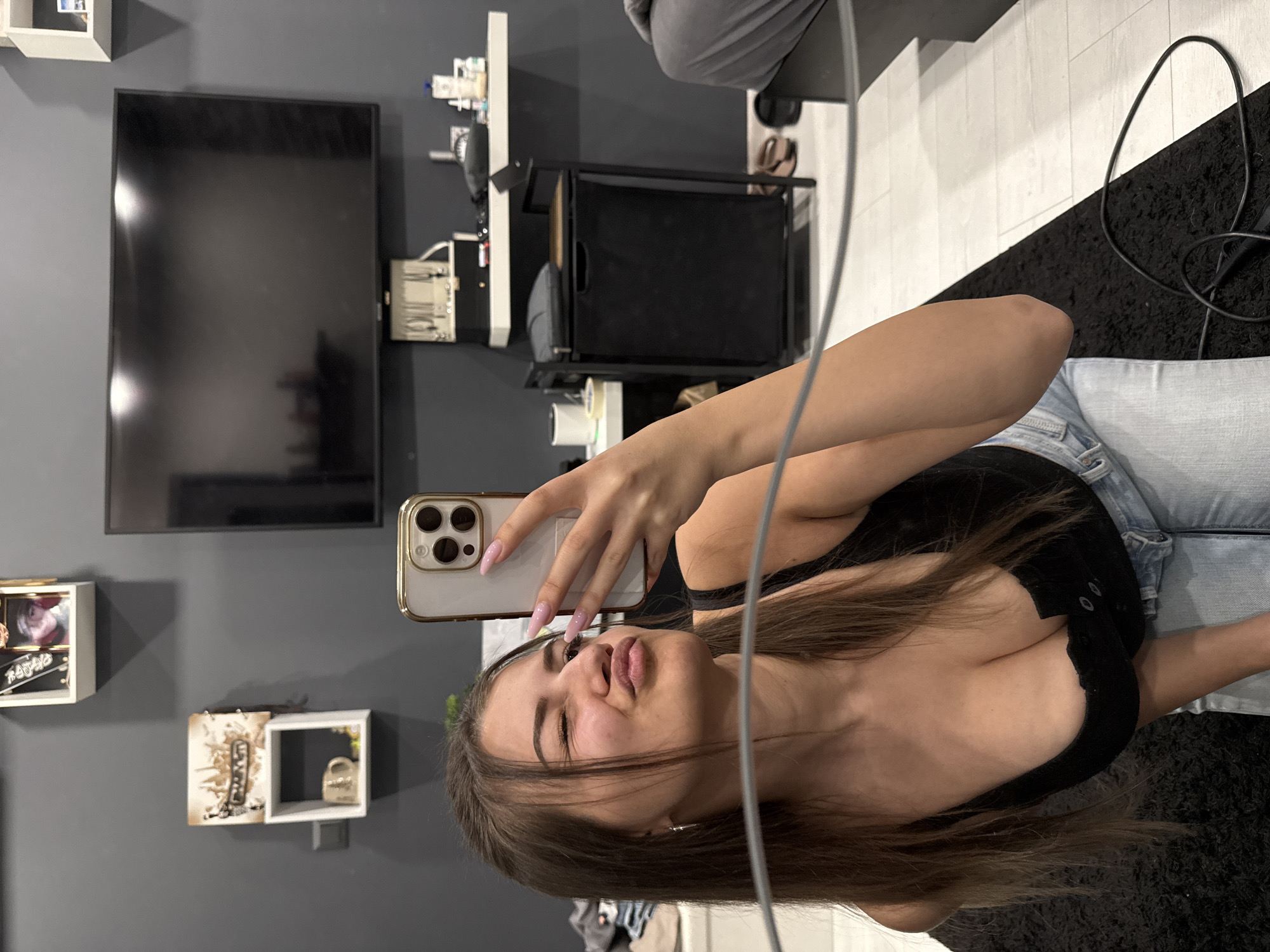 A young woman captures a selfie in a reflective surface, likely a mirror, in an indoor setting. The image is oriented with the woman's head to the left and her feet to the right, implying a 90-degree clockwise rotation from typical portrait orientation. She has long brown hair, is wearing a black top and light blue jeans, and holds a gold-edged smartphone with pink acrylic nails. Her expression is a playful pout or "duck face."
The background reveals a room with dark grey walls. To the upper left (when viewed as rotated), a black flat-screen television is mounted on the wall. To the right of the TV, a white shelf holds various bottles and small items. Below this, a black metal utility cart or trolley with what appears to be storage baskets is visible. Further to the right, a white tiled floor transitions into a black shaggy rug. A white cable runs vertically down the center-right of the frame. On the bottom left wall, two small white cube shelves display decorative items, including what looks like a small golden lock in one. The overall ambiance suggests a well-kept, contemporary indoor space, possibly a dressing area, salon, or bedroom. There is no visible text in the image. The location context provided is Sofia, Bulgaria.