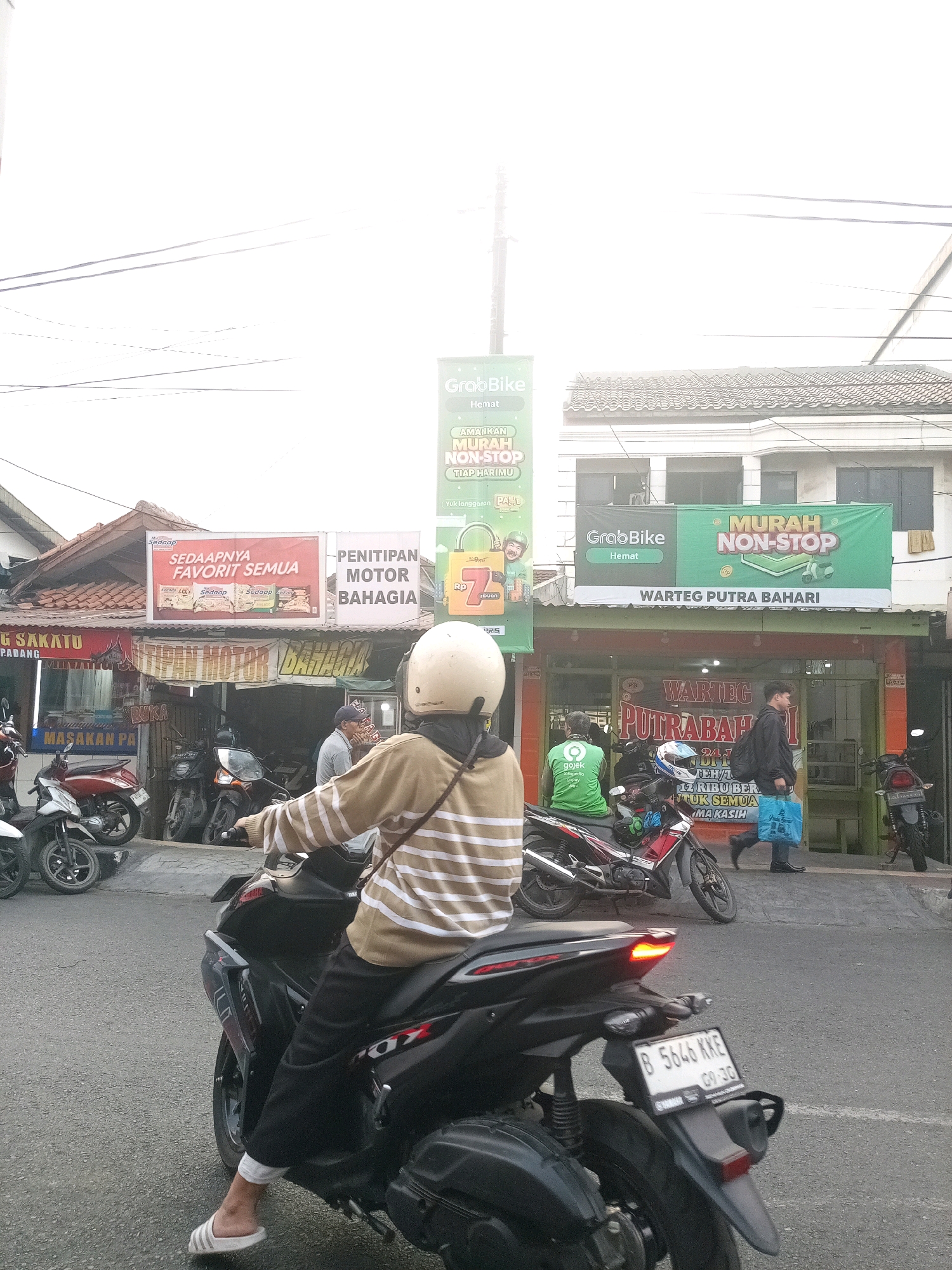 A street scene in Bekasi, Indonesia, depicts a pedestrian crossing in front of several small businesses and parked motorcycles. A person in a striped sweater and white helmet rides a black scooter in the foreground, their left foot clad in a white sandal. Behind them, a green Gojek rider waits on a motorcycle. The businesses include "WARTEG PUTRA BAHARI" and a motorcycle repair shop with a sign reading "PENITIPAN MOTOR BAHAGIA" and "BUKA". A large banner advertises "GRAB BIKE MURAH NON-STOP". Several parked motorcycles are visible, including red and black models. The sky is overcast, and electrical wires crisscross the frame. The license plate of the scooter in the foreground reads "B 5646 KKE".