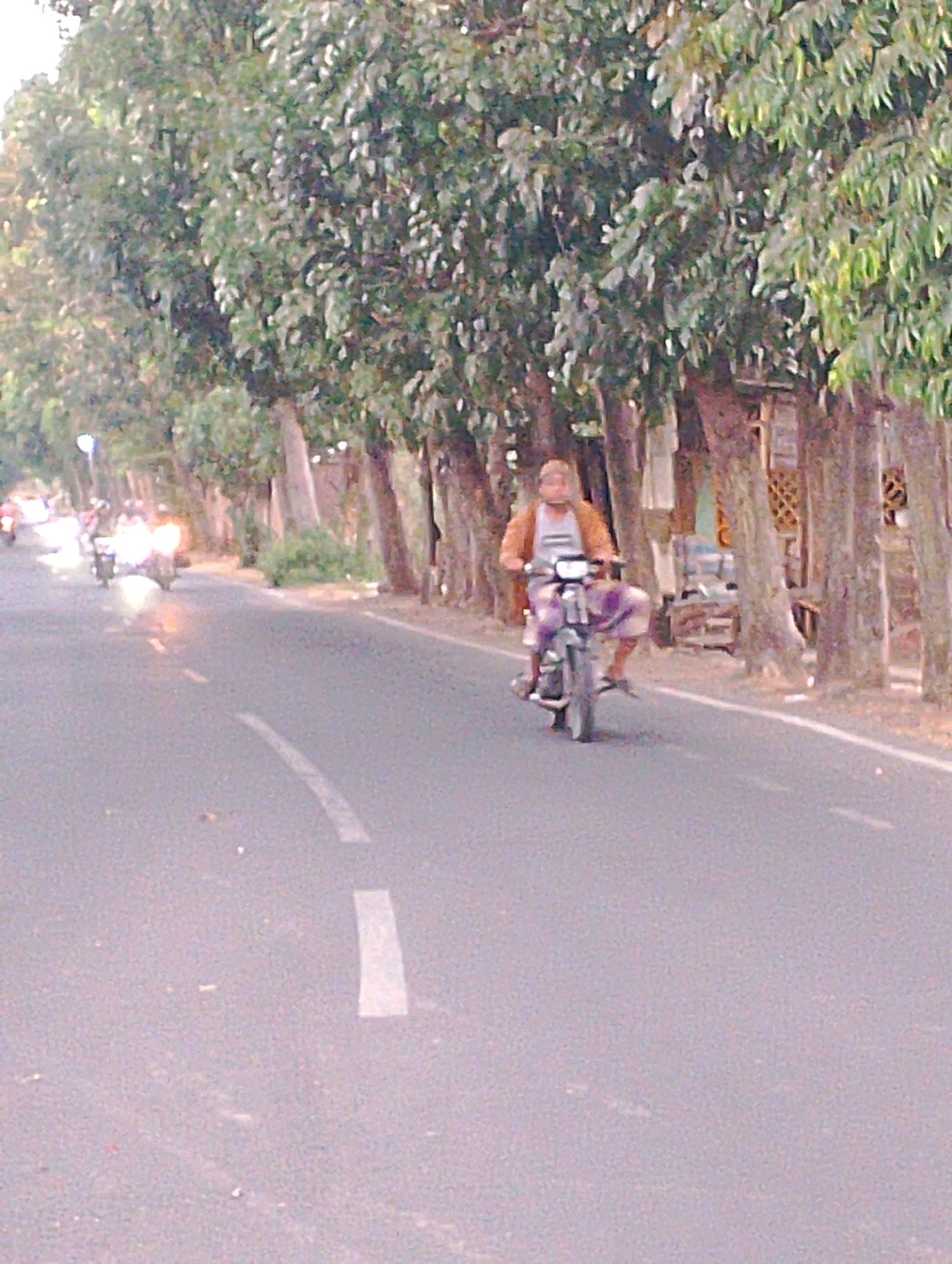 A low-resolution, outdoor, daytime, eye-level shot captures a man riding a motorcycle on a paved road lined with trees. The man is wearing a light-colored sleeveless shirt and an orange jacket. He is riding a dark-colored motorcycle and appears to be wearing patterned pants. The road has a dashed white line on the left side, and a solid white line on the right. In the background, to the left of the motorcycle, a line of motorcycles with illuminated headlights are visible, suggesting a procession or group of riders. The trees are tall and leafy, with some visible structures behind them on the right side of the road, possibly houses or market stalls with woven bamboo elements. The overall scene depicts a typical street scene in Probolinggo, Indonesia.