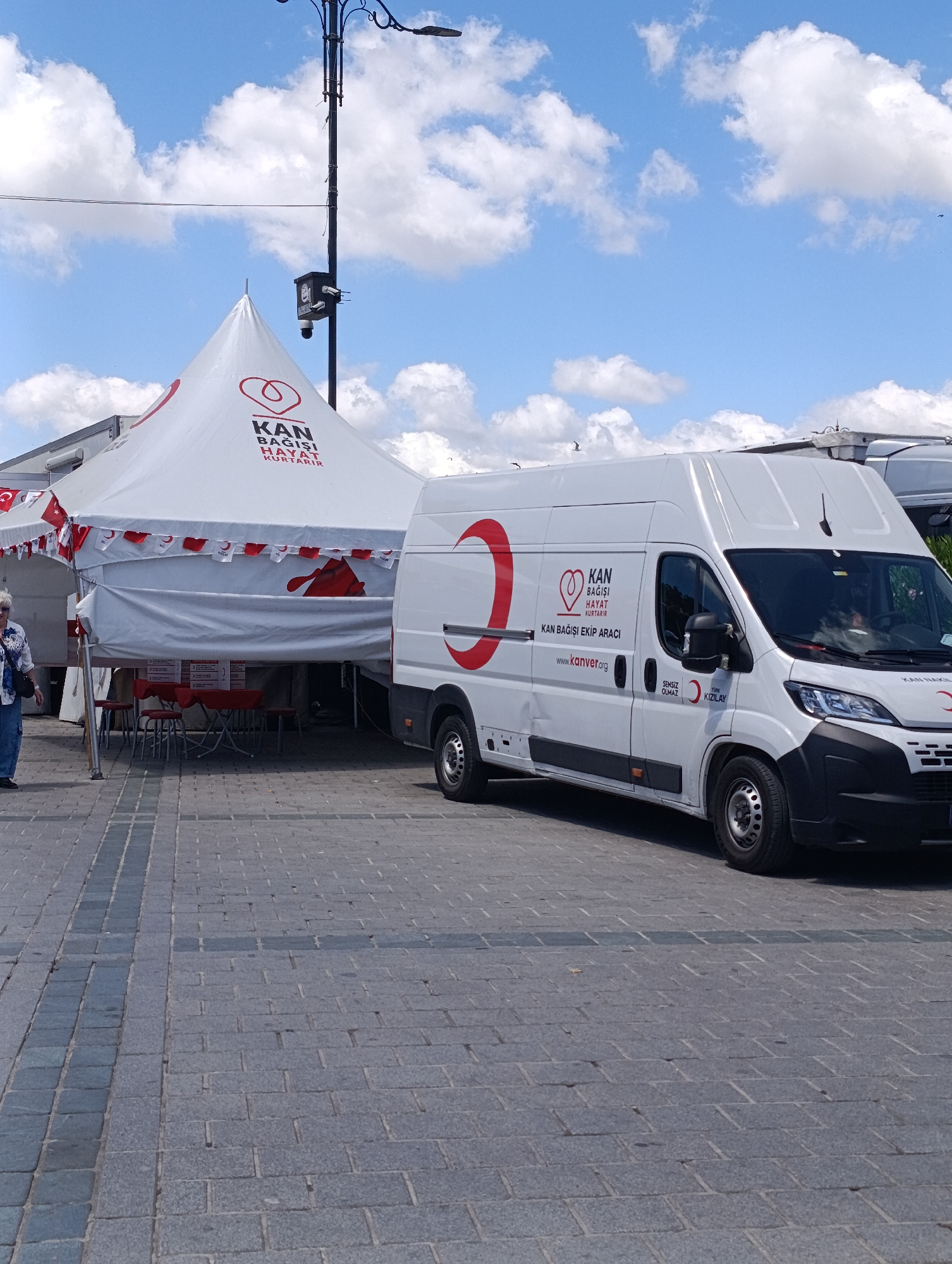 The image depicts a blood donation event set up in an urban outdoor plaza, likely in Istanbul, Turkey, given the Turkish text and the presence of Turkish flags. In the foreground, a white van with Turkish Red Crescent (Kızılay) branding is parked on a paved surface. The van has a large red crescent logo on its side and text that reads "KAN BAĞIŞI HAYAT KURTARIR" (Blood Donation Saves Lives) and "KAN BAĞIŞI EKİP ARACI" (Blood Donation Team Vehicle). Adjacent to the van is a large white tent, also adorned with Turkish Red Crescent symbols and text. Small Turkish flags are strung along the front of the tent. Inside the tent, red tables and stools are visible, suggesting a waiting or registration area. A woman is partially visible on the left side of the image, walking away from the camera. The ground is paved with rectangular stones, and a lamppost with a security camera attached stands to the left of the tent. The sky above is blue with scattered white clouds.
