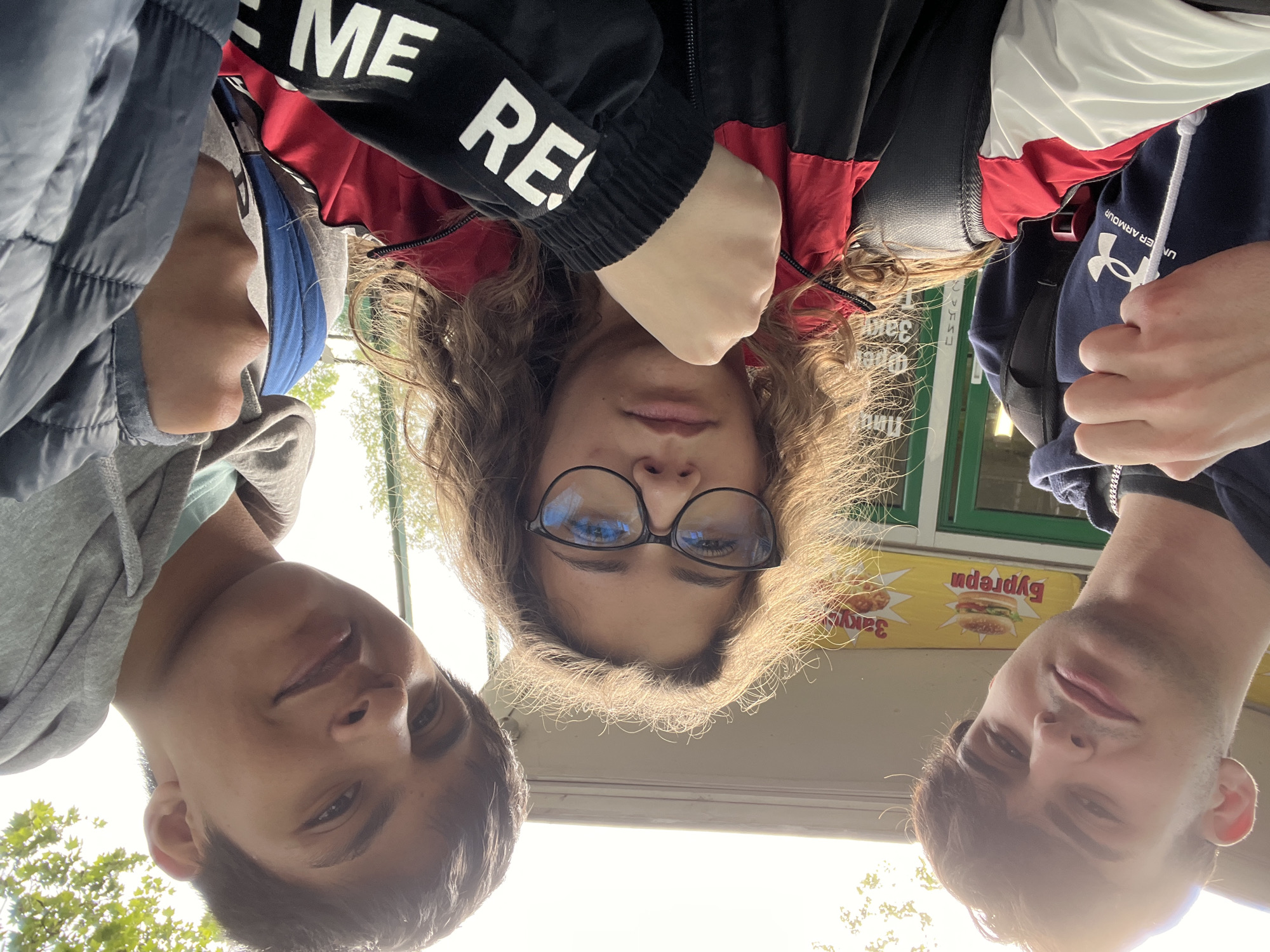 A low-angle shot captures three young individuals looking down at the camera, forming a triangular composition. The scene is outdoors during the daytime, likely late afternoon, with bright light coming from below and behind the subjects, creating some lens flare.
In the center, a young woman with long, curly light brown hair wears round glasses and a black, red, and white jacket. Part of a white word, "ME RE," is visible on her left sleeve. She looks directly at the camera with a neutral expression.
To her left, a young man with short dark hair is partially visible, wearing a gray hoodie and what appears to be a blue cap or hood under another layer. His face is slightly shaded.
To her right, another young man with short dark hair wears a dark blue hoodie that clearly displays the white "UNDER ARMOUR" logo. He also looks towards the camera with a neutral to slightly smiling expression.
The background includes a bright sky with green foliage from trees on the left. On the right, behind the subjects, is a building facade, possibly a kiosk or small shop, with a green-framed window. Two yellow signs are affixed to this building. The sign on the right features an image of a burger and the Cyrillic word "Бургер" (Burger). The sign on the left shows a fried chicken drumstick and the Cyrillic word "Закуски" (Snacks). The visible Cyrillic text suggests a location in a Slavic-speaking country, aligning with the provided context of Plovdiv, Bulgaria.