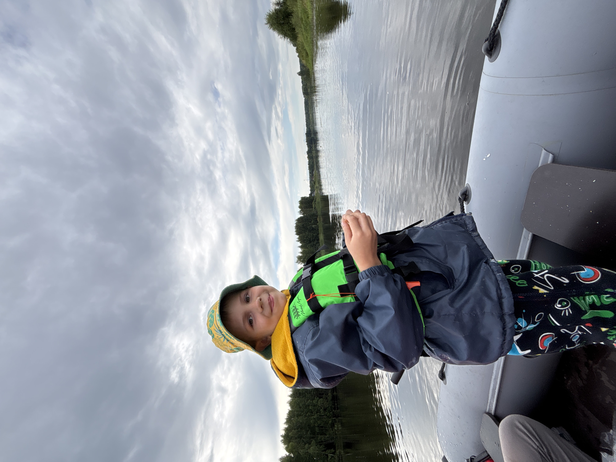 A young boy wearing a yellow and green patterned bucket hat, a blue jacket with a yellow hood, and a bright green life vest stands in an inflatable boat on a body of water. He is holding something small in his hands. The boat is positioned on a wide river or lake, with a tree-lined shore visible in the background. The sky is overcast with dramatic clouds. The scene appears to be in Anteplevo (Okhotinskoe S/P), Russia.