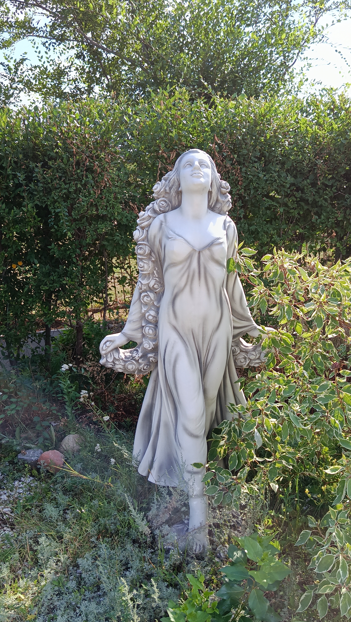 A life-sized, pale gray statue of a woman stands in a garden. The statue depicts a woman with her head tilted back and eyes looking upwards, framed by what appears to be sculpted roses intertwined with her hair. She wears a flowing, Grecian-style gown with a low neckline. Her arms are outstretched, with her hands open and palms facing upwards. The statue is positioned amongst dense green foliage and under a clear sky. To the right of the statue, a bushy plant with variegated green and white leaves is prominent. The ground around the statue is covered with various plants, grasses, and some small stones, one of which is reddish-brown. The overall scene suggests an outdoor garden setting. The location is identified as Kostanay, Kazakhstan.