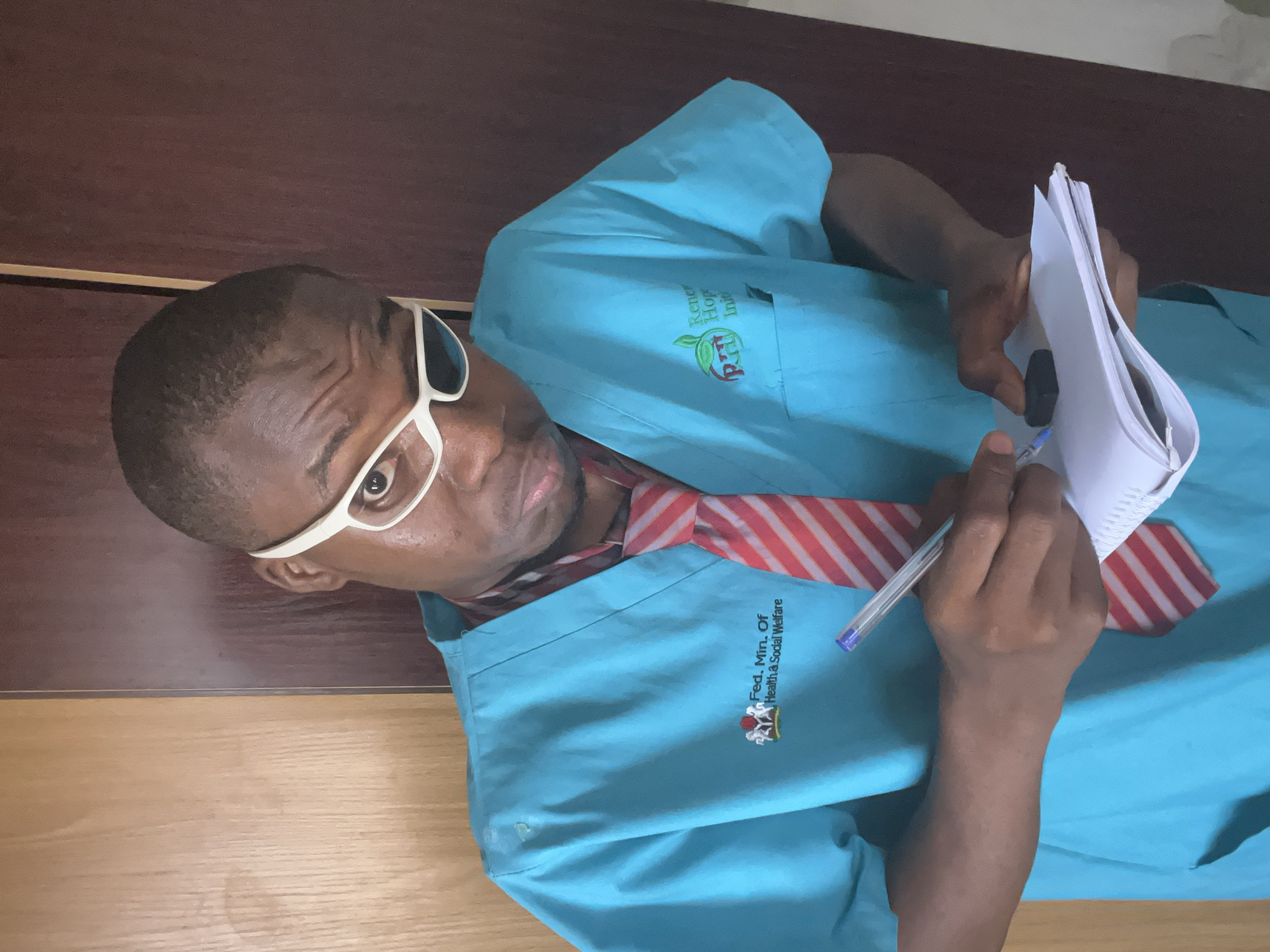 The image, rotated 90 degrees clockwise, depicts a man indoors, likely in Minna, Nigeria. He has dark skin, short dark hair, and wears white-framed glasses, a light blue scrub top, and a red and white striped tie. He looks directly at the camera with a neutral, focused expression.
In his right hand, he holds an open white notebook or folder with a black binding, while his left hand holds a blue pen, poised as if ready to write or having just written.
His light blue scrub top features two embroidered logos. On his right side (viewer's left), a green leaf design accompanies the partial text "Renewed Hope Initia" and the acronym "RHI". On his left side (viewer's right), the text "Fed. Min. Of Health & Social Welfare" is clearly visible, along with a logo of two intertwined figures.
The background consists of a dark brown wooden surface above and a lighter, horizontally-grained wooden surface below, indicating an office or clinical setting. The visible text suggests his affiliation with a governmental health and social welfare organization.