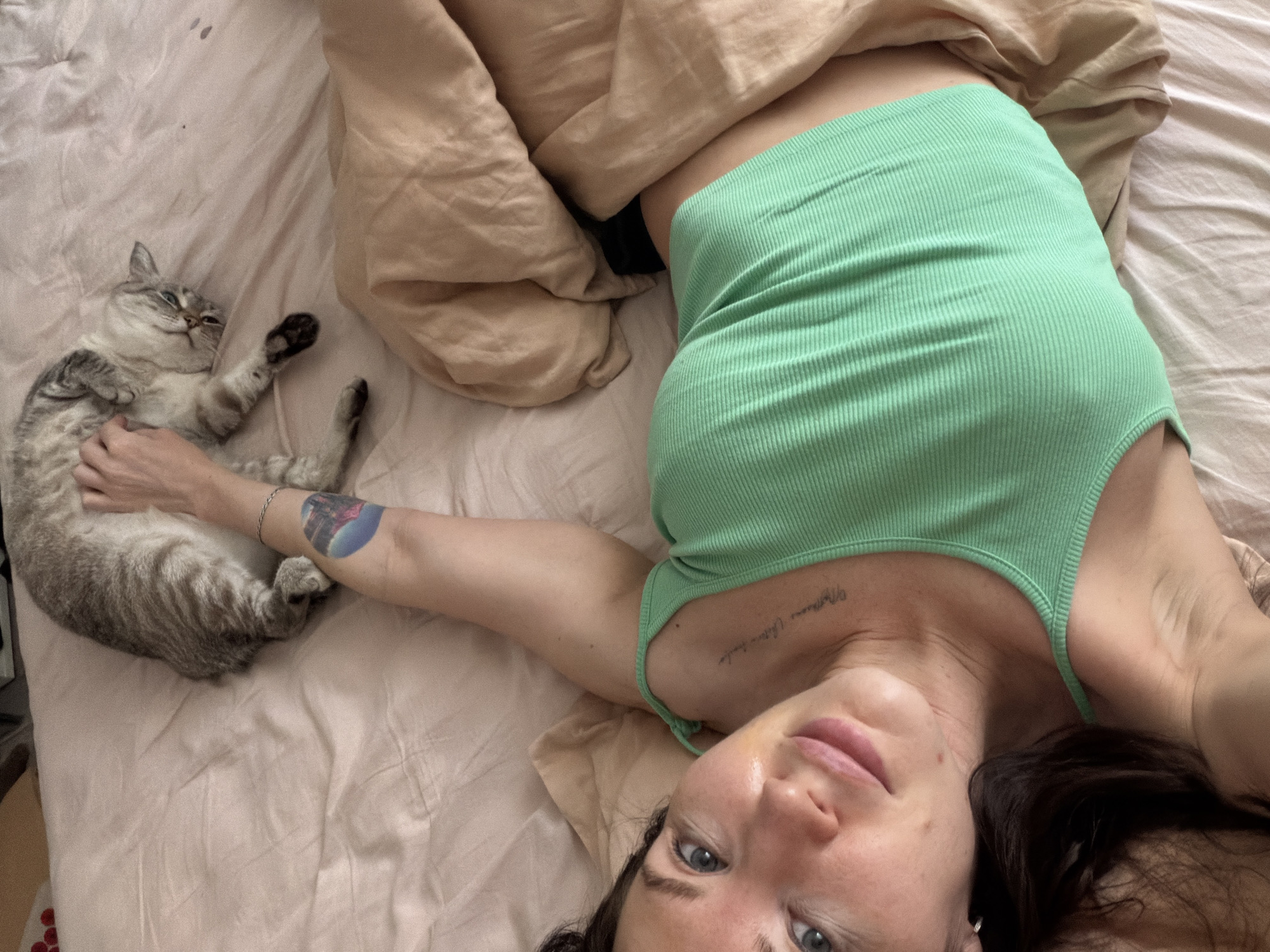 A woman lies on a bed, with a cat beside her. The woman is fair-skinned and has dark hair, wearing a green tank top. She is looking at the camera. Her right arm rests on the cat's back, which is gray and tabby-striped. The bed has light beige sheets and a rumpled beige blanket. The location is possibly Perm, Russia.